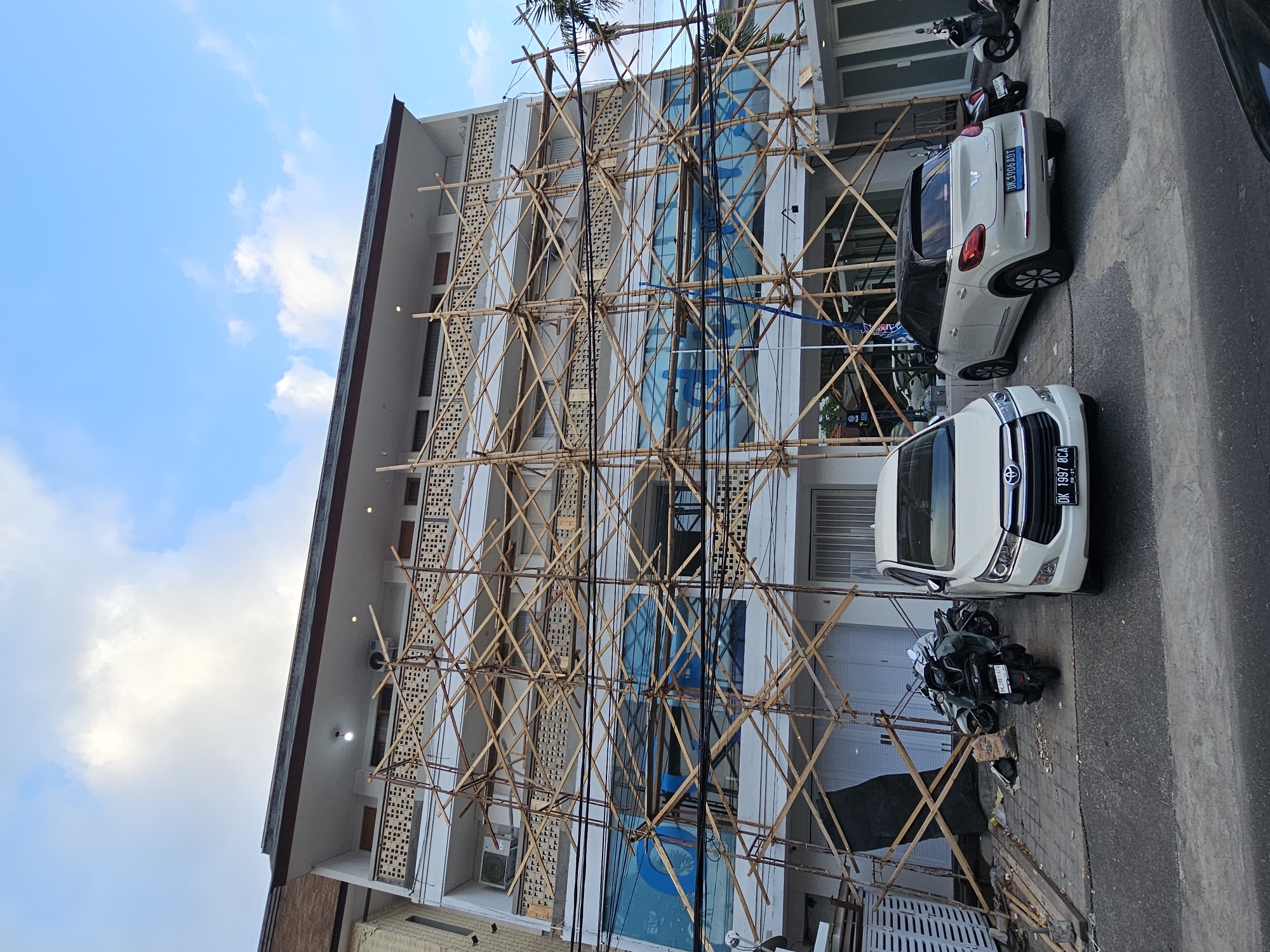 The image shows a building under construction in Denpasar, Indonesia. Bamboo scaffolding covers the facade. Parked on the street in front of the building are two cars, a white Toyota and a light-colored Fiat. Also parked nearby is a motorcycle. The sky is visible in the upper left corner with scattered clouds.