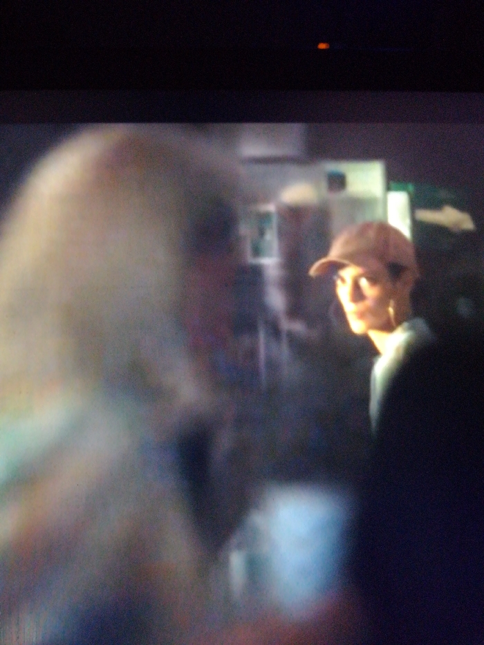 A blurry image shows two individuals, one out of focus in the foreground with fair hair. In the background, a person wearing a brown baseball cap looks directly at the camera. The scene is dimly lit, obscuring details of their clothing and the environment. There's a white sign with an arrow, which suggests an indoor location possibly.