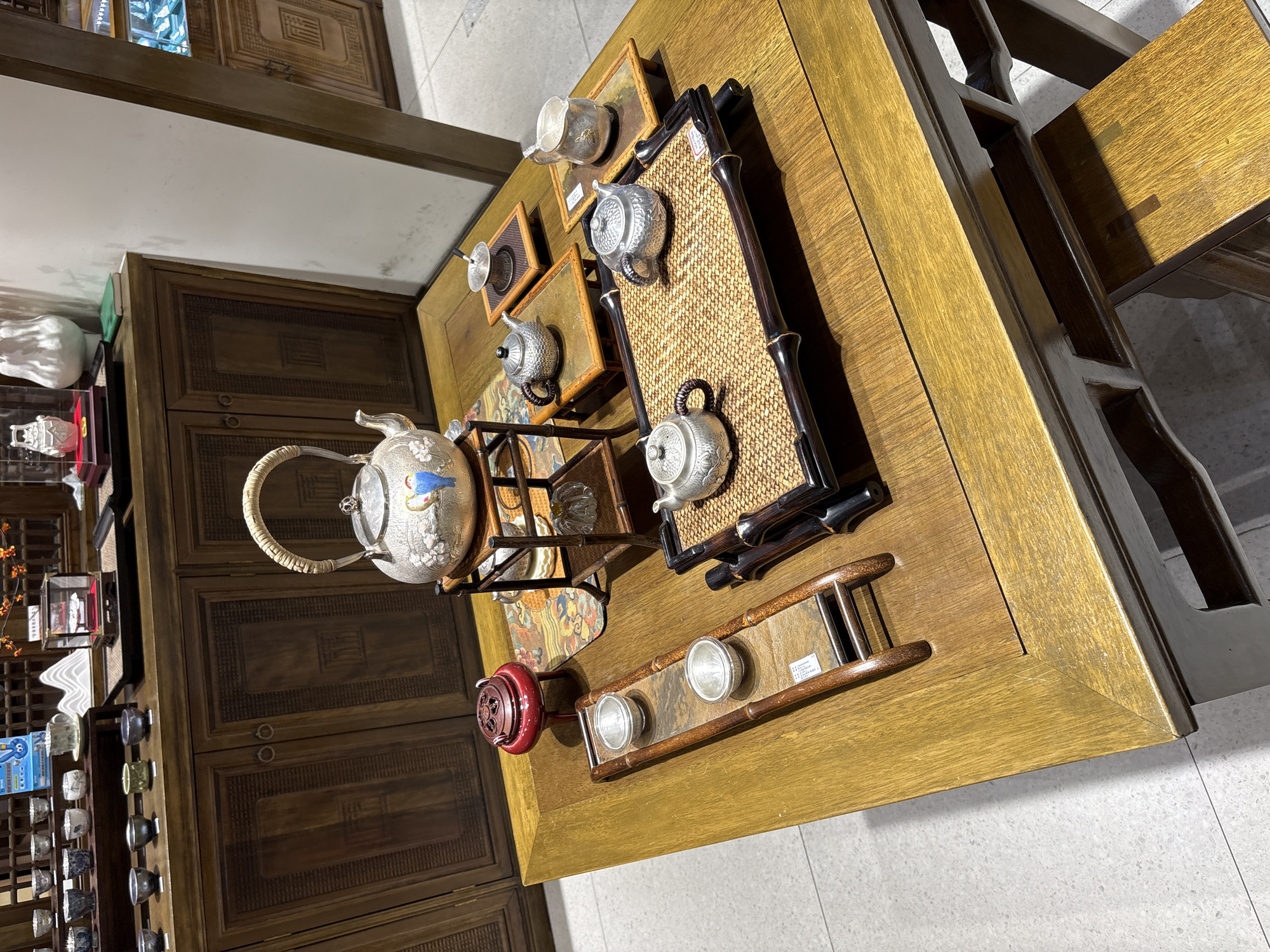 An ornate, silver teapot adorned with painted birds sits atop a multi-tiered stand on a polished wooden table. Arranged around the teapot are several smaller silver teacups, a silver creamer, and a silver sugar bowl, some resting on individual wooden trays. A red incense burner with intricate carvings is positioned to the left of the main display, on a long, narrow wooden tray holding two silver cups. The table itself is large and made of warm-toned wood, with visible grain patterns. Behind the table, a dark wooden cabinet with carved doors serves as a backdrop, displaying rows of smaller silver cups on shelves. The scene appears to be set in an indoor establishment, possibly a tea shop or a display area within a retail space. The lighting is soft and ambient, creating a warm and inviting atmosphere. The overall impression is one of elegance and tradition, highlighting artisanal craftsmanship in tea ware.