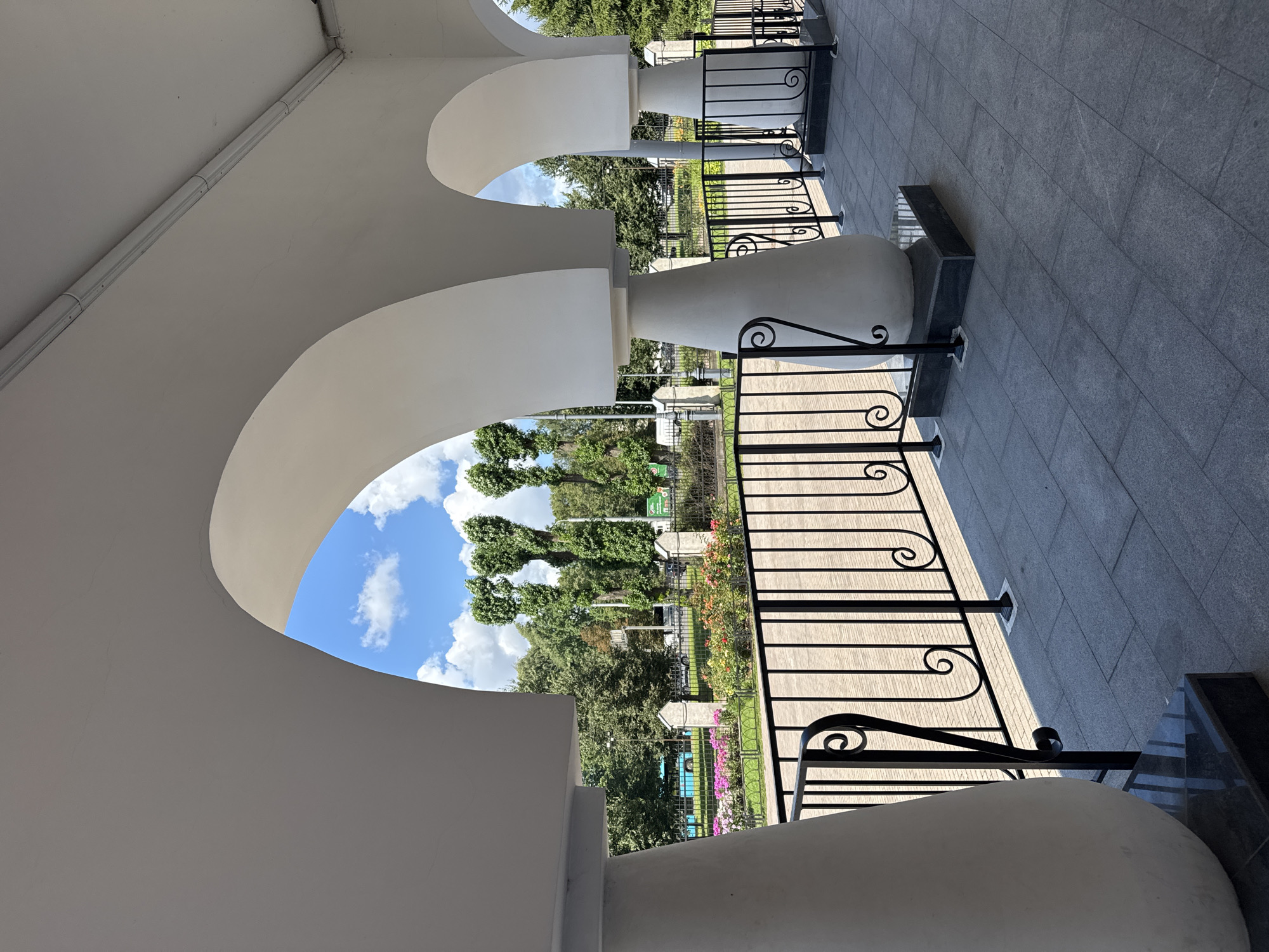 The image shows a covered walkway with white arched ceilings supported by thick, bulbous white pillars. A black metal railing with decorative scrollwork runs along the edge of the walkway. Beyond the railing, a paved path is visible, bordered by a flower bed with colorful flowers. Further in the background, a black metal fence encloses a green lawn and several trees. Among the trees, tall, slender trees with distinctive rounded tops are noticeable. Beyond the trees, a blue bus and other vehicles are visible on a road, suggesting an urban or park setting. The sky is blue with scattered white clouds. The overall impression is that of a well-maintained outdoor space, possibly part of a park, museum, or public building in Saint Petersburg, Russia.