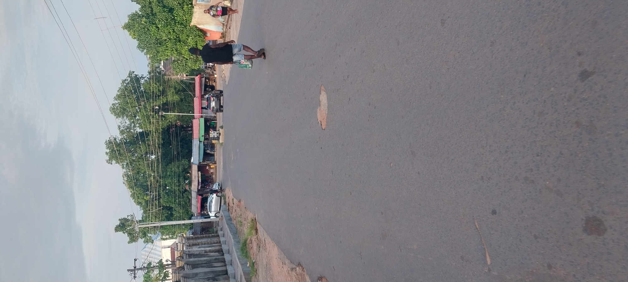 A straight, asphalt road stretches into the distance in Onitsha, Nigeria. The road has some patches of disrepair, with a noticeable pothole in the middle of the frame. To the left, a concrete barrier with decorative pillars lines the side of the road, followed by a low wall and some vegetation. On the right side of the road, a woman walks away from the camera, carrying a bag. Further down the road, a man walks towards the camera, also carrying a bag. A dark-colored SUV is parked on the right side of the road, and a silver car is parked on the left. In the background, a row of small shops with colorful awnings are visible, backed by lush green trees under a cloudy sky. Power lines are strung across the scene.
