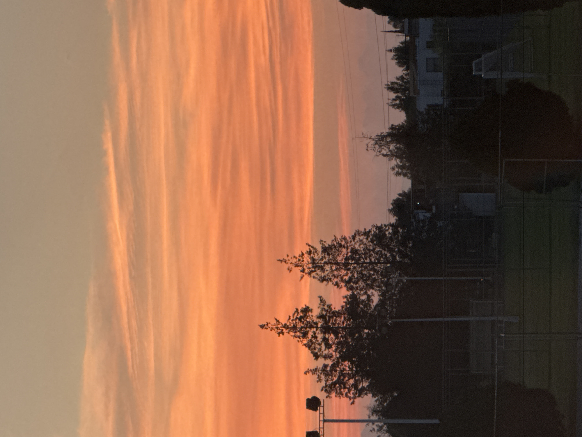 The image shows a landscape at twilight in Heroica Puebla de Zaragoza, Mexico. A vibrant, orange-colored sky with linear cloud formations dominates the left side. Silhouetted trees, possibly a park, are visible in the middle of the image. Light poles are interspersed among the trees. A building and a fenced-in area possibly used for a sporting activity can be discerned on the right side of the image. Overall, the setting appears to be a residential area bordering a park at dusk.