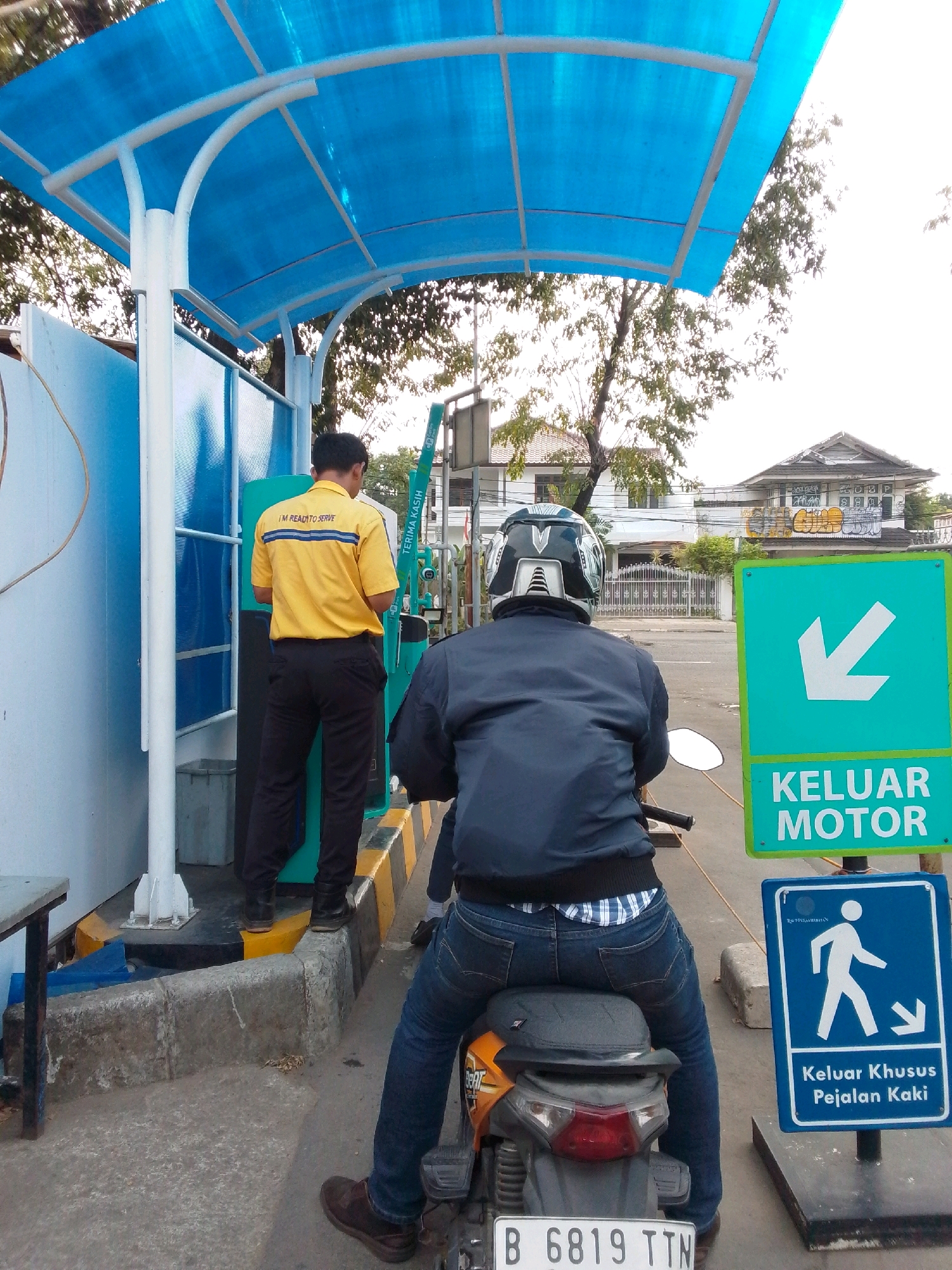 This is a medium-shot, slightly low-angle photograph taken outdoors in East Jakarta, Indonesia. The scene depicts an outdoor payment or service area, likely for vehicles. In the foreground, a person is seated on a motorcycle, facing away from the camera. They are wearing a dark jacket, blue jeans, and a helmet. The motorcycle has a license plate that reads "B 6819 TTN". To the right of the motorcycle, there are two signs. The top sign is green with a white arrow pointing left and the text "KELUAR MOTOR". Below this is a blue sign with a white silhouette of a person walking and an arrow pointing down, along with the text "Keluar Khusus Pejalan Kaki". In the mid-ground, a person wearing a yellow shirt with "I'M READY TO SERVE" printed on it and dark pants is standing near a green payment machine. The person in the yellow shirt appears to be interacting with the machine. Behind them, a blue and white structure with a blue canopy provides shelter. In the background, trees and residential buildings are visible. The overall atmosphere suggests a transaction or service point within a bustling urban environment.