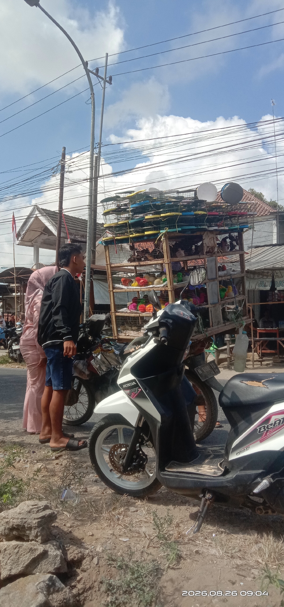 The image depicts a street scene in Pakong, Indonesia, on a sunny day. In the foreground, two motorcycles are parked on a dirt and gravel shoulder next to a paved road. The motorcycle on the right is white and black with "HONDA" and "FI" visible on its side. The motorcycle behind it is partially visible.
Two people are standing on the left side of the road. One is wearing a long, light pink garment and a headscarf, and the other is wearing a black jacket and blue shorts.
Behind the motorcycles, a wooden cart is loaded with items. The lower shelves of the cart contain brightly colored, spherical objects, possibly toys or decorative items. The upper shelves are stacked with numerous green, yellow, and blue cages or containers, some of which appear to have lids.
In the background, there are buildings with tiled roofs, electrical wires, and a utility pole. A red and white flag, possibly the Indonesian flag, is visible on a pole near a building with a white facade. The sky is blue with scattered white clouds. The overall scene suggests a marketplace or roadside vendor stall.