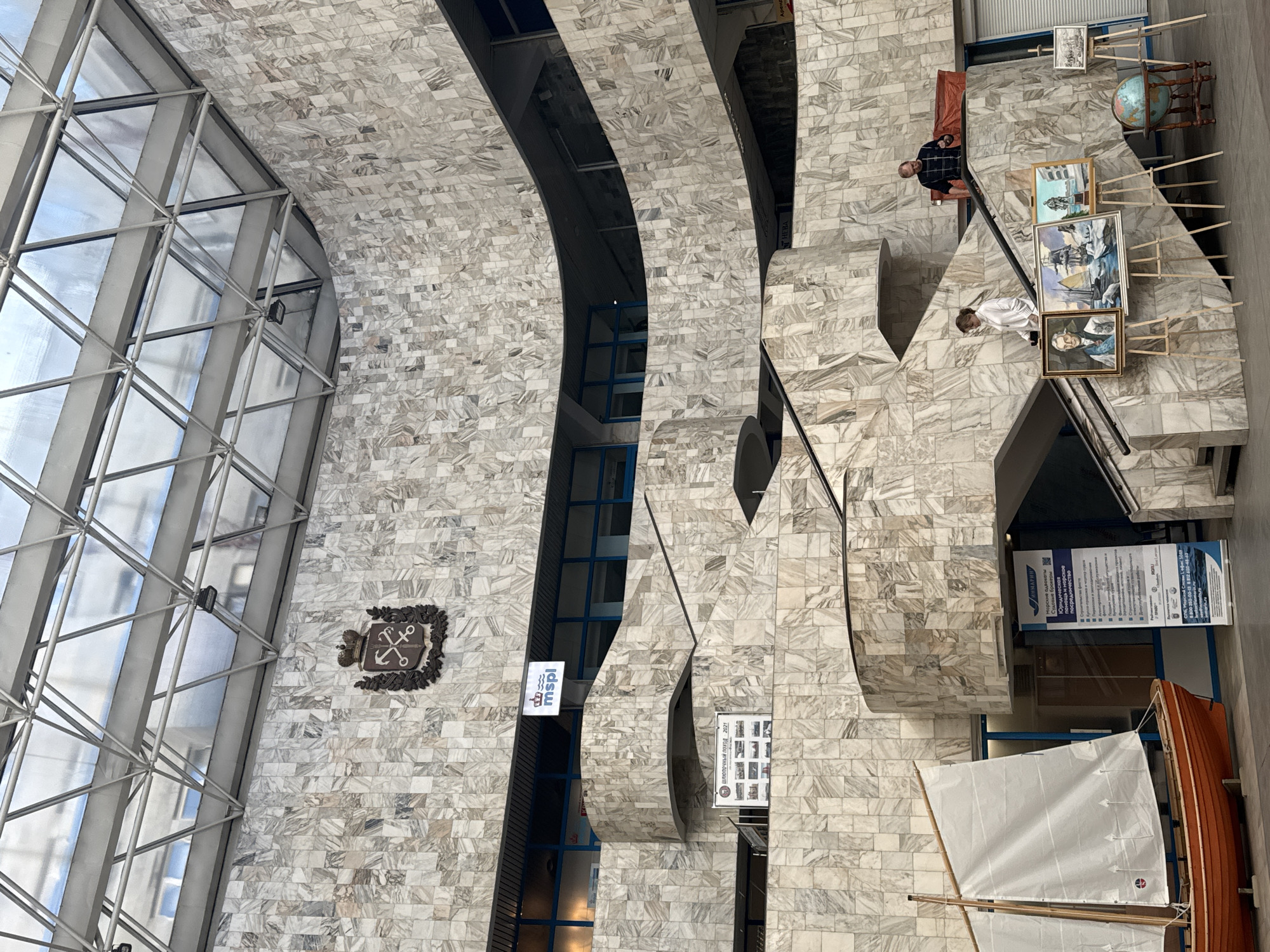 The interior of a building, possibly a maritime museum or institution, features an atrium wall made of marble-like stone tiles. The wall is punctuated by sections with windows and curved architectural elements. A large window extends from the left side of the image, allowing natural light to fill the space. A maritime emblem is affixed to the wall next to the window.
On the right side of the image, two individuals are present on an upper level. One is seated and wearing a plaid shirt, while the other is standing and appears to be wearing a white shirt or jacket. A display of framed artwork is positioned on the same level, along with a globe and other items on stands. Below, there's a small model sailboat with a white sail. A sign with Russian text is visible near the boat. The location is likely in Saint Petersburg, Russia, suggested by the Russian text on the signs.