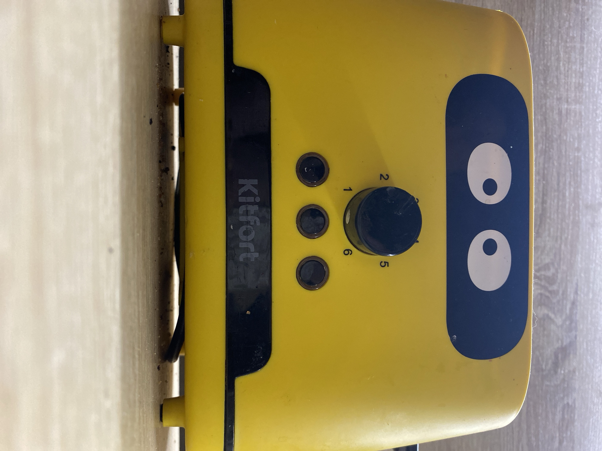 A yellow electronic appliance, likely a toaster, is depicted in a close-up, rotated image. The main body of the appliance is a bright yellow, featuring a black control panel down its center. The brand name "Kitfort" is visible in white lettering on this black panel, oriented vertically from bottom to top in the current image's rotation.
To the right of the brand name, three small, dark circular buttons are arranged vertically. Above these, a larger black circular knob is present, marked with numbers "1, 2, 5, 6" around its perimeter, suggesting a browning or timer setting.
A prominent design feature on the right side of the appliance is a large, dark blue rounded rectangle containing two white, oval-shaped "eyes" with black pupils, giving the device a cartoonish, friendly appearance.
The appliance is positioned against two different surfaces. To its left, a light-colored, possibly wooden, surface is visible, showing some dust and debris in the crevice where it meets the appliance. A black cable is also partially visible here. To its right, a lighter, textured surface, possibly a countertop or floor, can be seen. The item is stationary, with no activity taking place. The overall impression is an indoor setting, consistent with a kitchen in Chaikovsky, Russia.