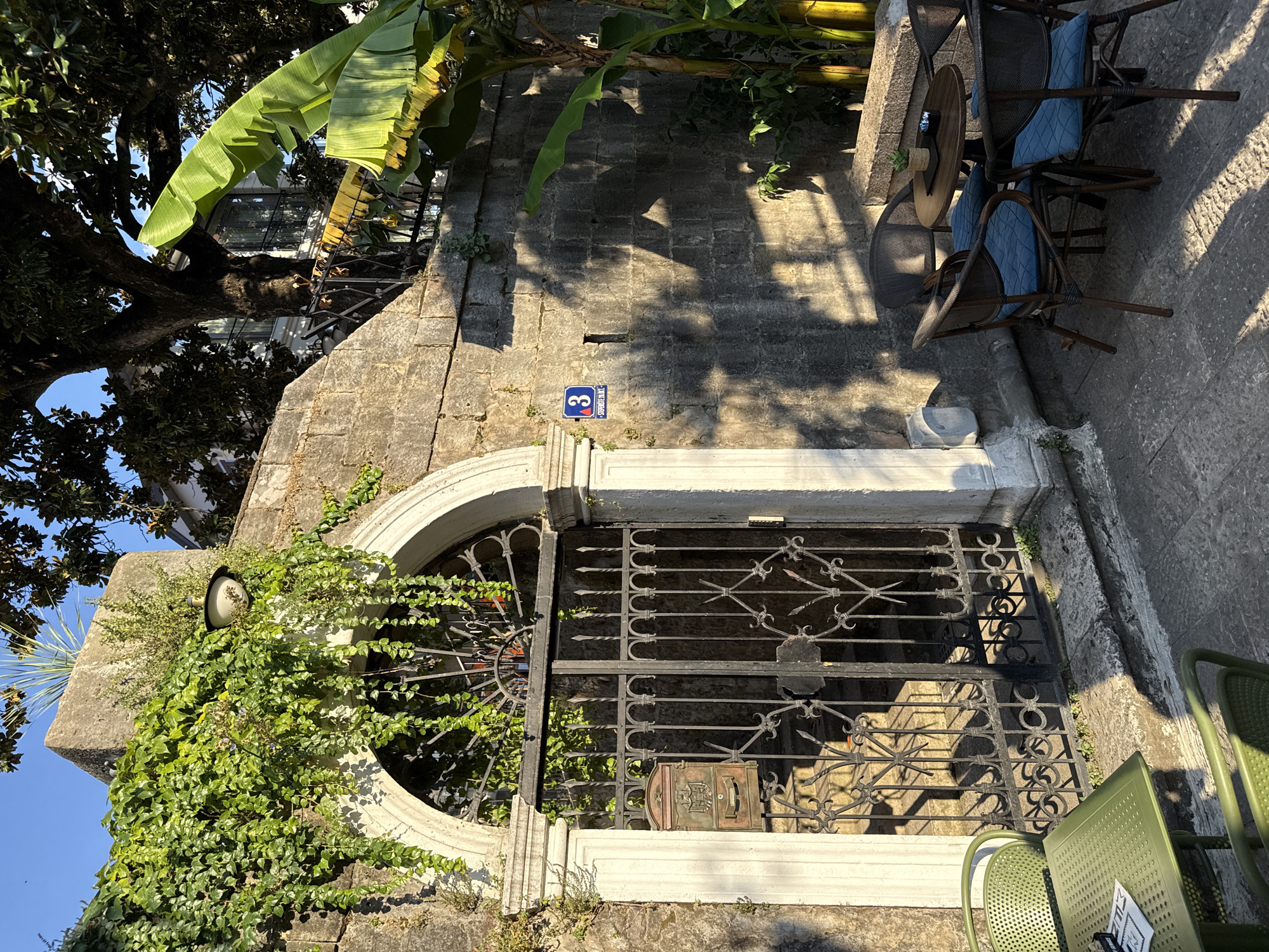 A low-angle, medium shot captures an outdoor seating area and an ornate metal gate in Herceg Novi, Montenegro, bathed in warm, late afternoon sunlight. The scene is dominated by a textured, stone wall with patches of vibrant green ivy climbing its surface. A white, arched entryway frames the dark metal gate, which is adorned with intricate scrollwork and topped with a decorative semi-circular iron element. A vintage-style mailbox is affixed to the left side of the gate. Above the gate, a white outdoor lamp is mounted on the stone wall. To the right of the gate, a blue and white street sign with the number "3" and an arrow is visible.
In the foreground to the left, a green metal table with a perforated top is set with what appears to be a menu or leaflet. A matching green metal chair is partially visible to the left of the table. To the right of the gate, a small outdoor seating arrangement consists of a round, dark wooden table with four woven chairs, each fitted with a plush blue cushion. A small potted plant and an ashtray sit on the table.
Lush vegetation, including large banana leaves and mature trees with dense foliage, adds a tropical feel to the background. The sunlight casts long, dramatic shadows across the stone wall and the courtyard floor, indicating the time of day. The overall impression is one of tranquil, European charm.