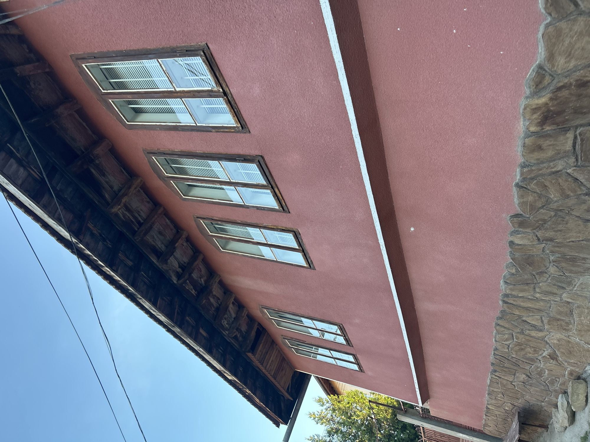 An exterior view of a two-story building, possibly a house or a small business, is captured from a low angle, looking upwards. The building has a reddish-pink stucco exterior, a pitched wooden roof with exposed beams, and several tall, narrow windows. These windows have wooden frames and are divided into two panes, with white blinds visible inside. The roofline extends out over the facade, supported by visible wooden rafters. A white horizontal band runs across the middle of the building's facade, separating the upper section from the lower section which is clad in rough-hewn stone. The stone base is irregular in shape and color, ranging from light brown to dark grey. To the left, a section of a fence and some trees are visible, suggesting a semi-rural or suburban setting. Electrical wires are strung across the top of the frame against a clear blue sky. The overall impression is of an older, possibly rustic, structure.