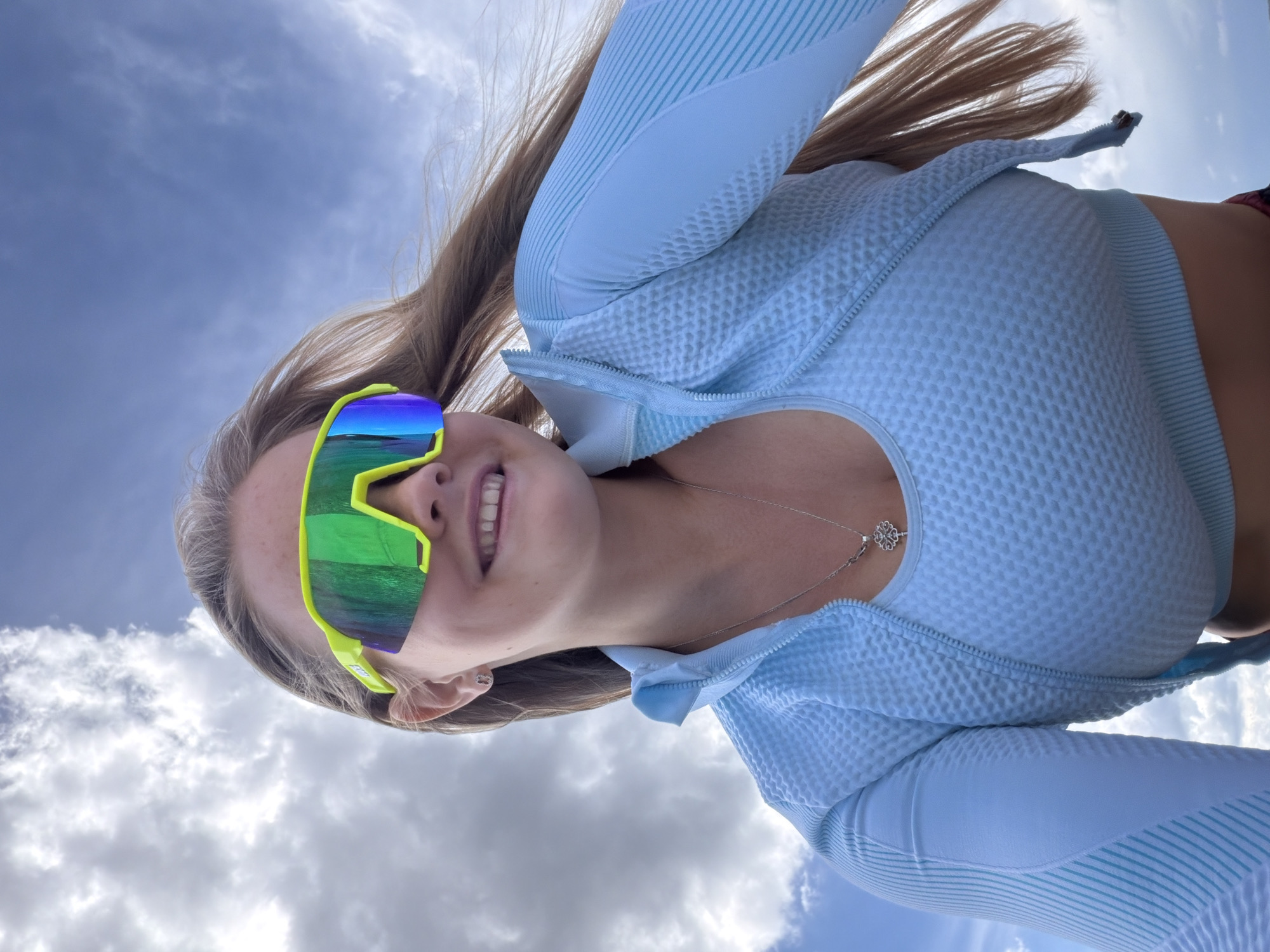 A young woman with blonde hair, wearing a light blue zip-up athletic top and bright green and yellow sports sunglasses, smiles broadly at the camera. She has a delicate silver necklace with a clover-shaped pendant. Her hair is pulled back and streams upward, suggesting movement or wind. The background is a vast expanse of sky with scattered clouds, indicating an outdoor setting. The image is shot from a low angle, looking up at the woman. The lighting suggests it is daytime, likely sunny given the brightness of the sky and the reflections on her sunglasses. The overall mood is cheerful and energetic.