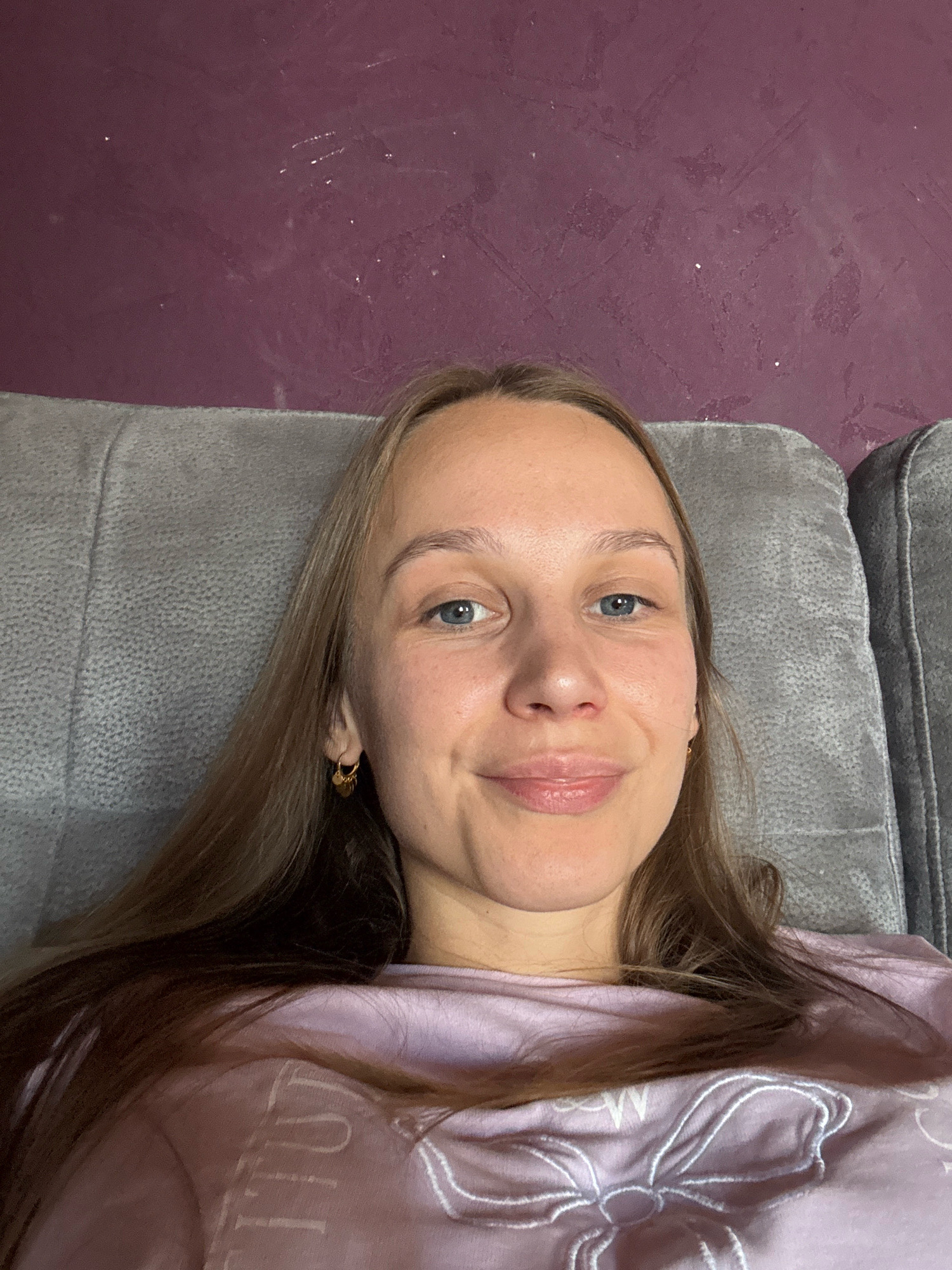 A young Caucasian woman with long, straight blonde hair is lying on a grey sofa. She is wearing a light pink t-shirt with a white graphic design on it. She has blue eyes, fair skin, and is smiling slightly. The background is a textured purple wall. The image appears to be a selfie taken indoors.
