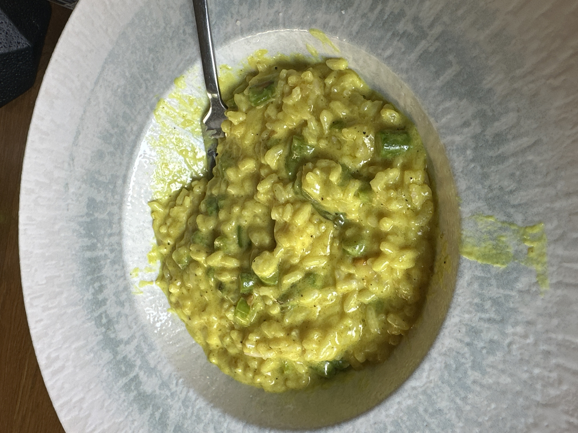 A close-up, overhead shot shows a plate of risotto in Sofia, Bulgaria. The risotto, yellow in color, is topped with what appear to be green vegetables, possibly asparagus or green onions. A silver fork rests within the risotto on the left side of the plate. The plate itself is round with a grey and white mottled pattern. The table surface is visible on the left, adjacent to the plate. A dark, patterned object is visible in the upper left corner of the frame.