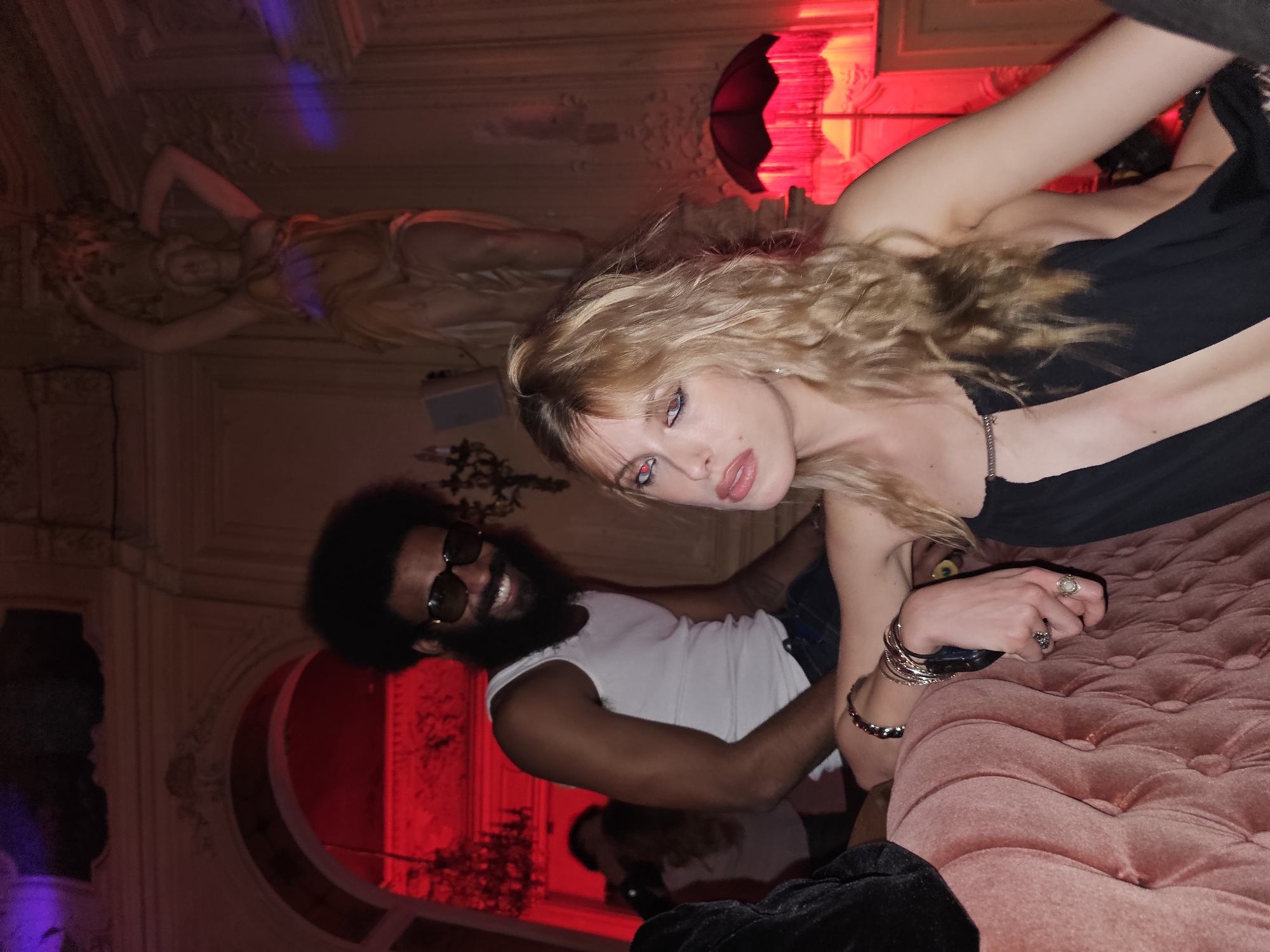 The photograph shows a Caucasian woman with blonde, wavy hair and pale skin, and an African-American man with a beard and sunglasses in an ornate interior. The woman is in the foreground wearing a black top, her hand adorned with bracelets and rings rests on a pink tufted cushion. The man is wearing a white t-shirt. The interior features red lighting and an overhead statue. The presence of elaborate architectural details suggests the interior is in Paris, France. The camera angle and facial expressions suggest a candid or posed snapshot, rather than a professional portrait.