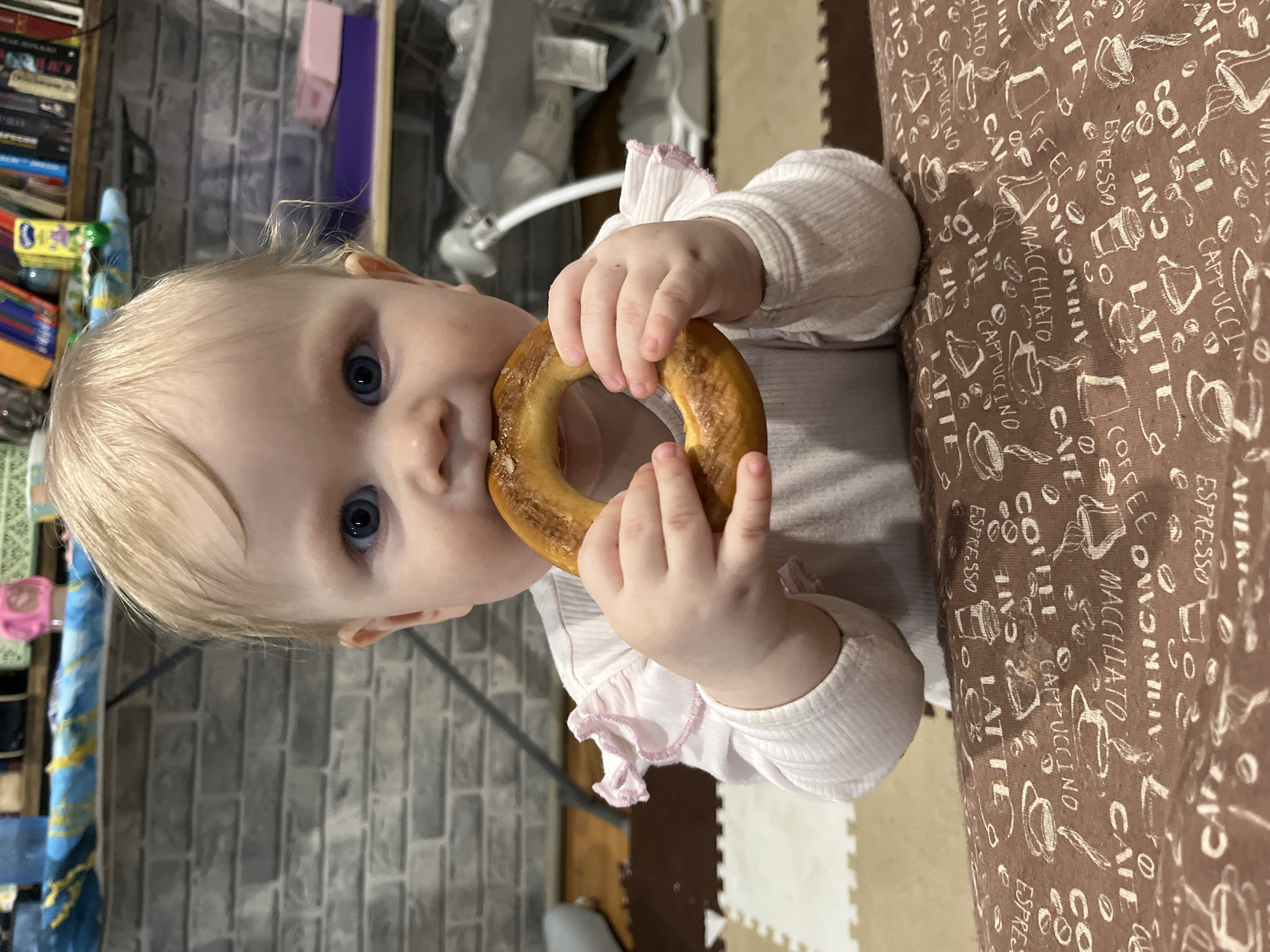 A close-up shot depicts a baby with blonde hair and blue eyes holding a ring-shaped baked good, possibly a bagel or bread ring. The baby is facing the camera and partially obscured by the baked good, which is held in both hands. The baby is wearing a long-sleeved white shirt with pink frill details. The background shows a gray brick wall and a shelf with books and other objects. A table or surface covered with a brown patterned cloth featuring various coffee-related terms and illustrations is visible on the right side. The playmat with puzzle pieces that are off-white and brown is visible behind the table. The image composition centers on the baby and the baked good. It is unlikely that the location of Vesyoloye, Russia can be determined from the image alone.