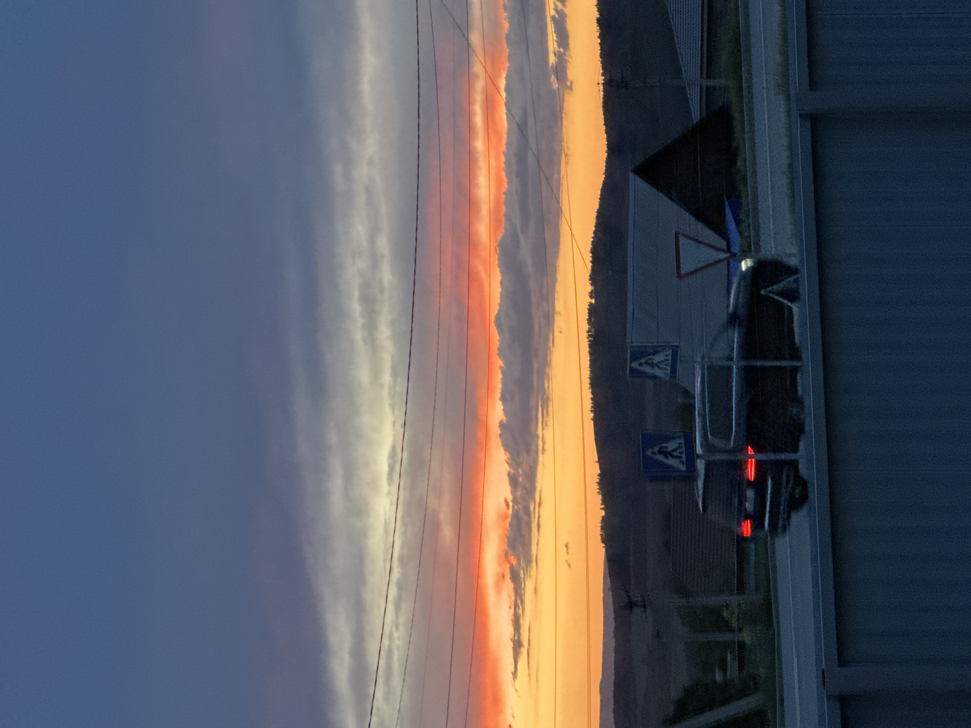 Captured in Uchaly, Russia, the image portrays a vertical scene showing a sky gradient from deep blue to vivid orange. A bank of grey-white clouds runs vertically along the left edge of the frame, transitioning into orange-tinted clouds. Power lines intersect the sky. The lower right portion of the image depicts roadside infrastructure including a building, a road, a triangular warning sign, and pedestrian crossing signs. Reflective tail lights suggest a vehicle is nearby.