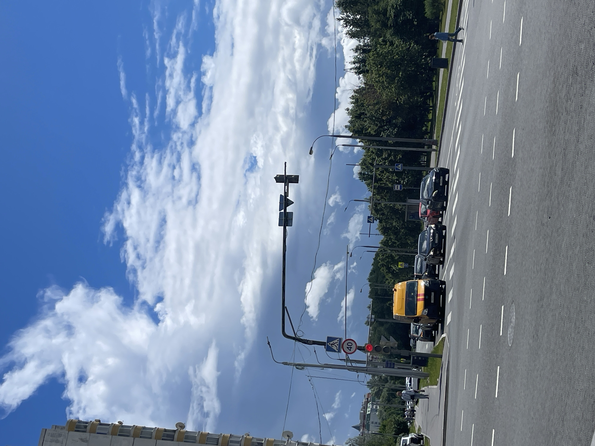 The image depicts a street scene in Moscow, Russia, on a partly cloudy day. The foreground shows a wide asphalt road with white dashed lane markings and a pedestrian crossing. In the middle ground, several cars, including a yellow van with red stripes, are stopped at a traffic light. A pedestrian is visible on the sidewalk to the left, and another person is crossing the street on the right. Traffic lights and signs are mounted on poles. A pedestrian crossing sign with a figure and a 40 km/h speed limit sign are visible. In the background, a tall, light-colored apartment building stands on the left, and lush green trees line the right side of the road. The sky is a vibrant blue with scattered white and gray clouds, some of which appear dark and heavy, suggesting the possibility of rain. The overall impression is of an urban environment with active traffic and pedestrian movement.