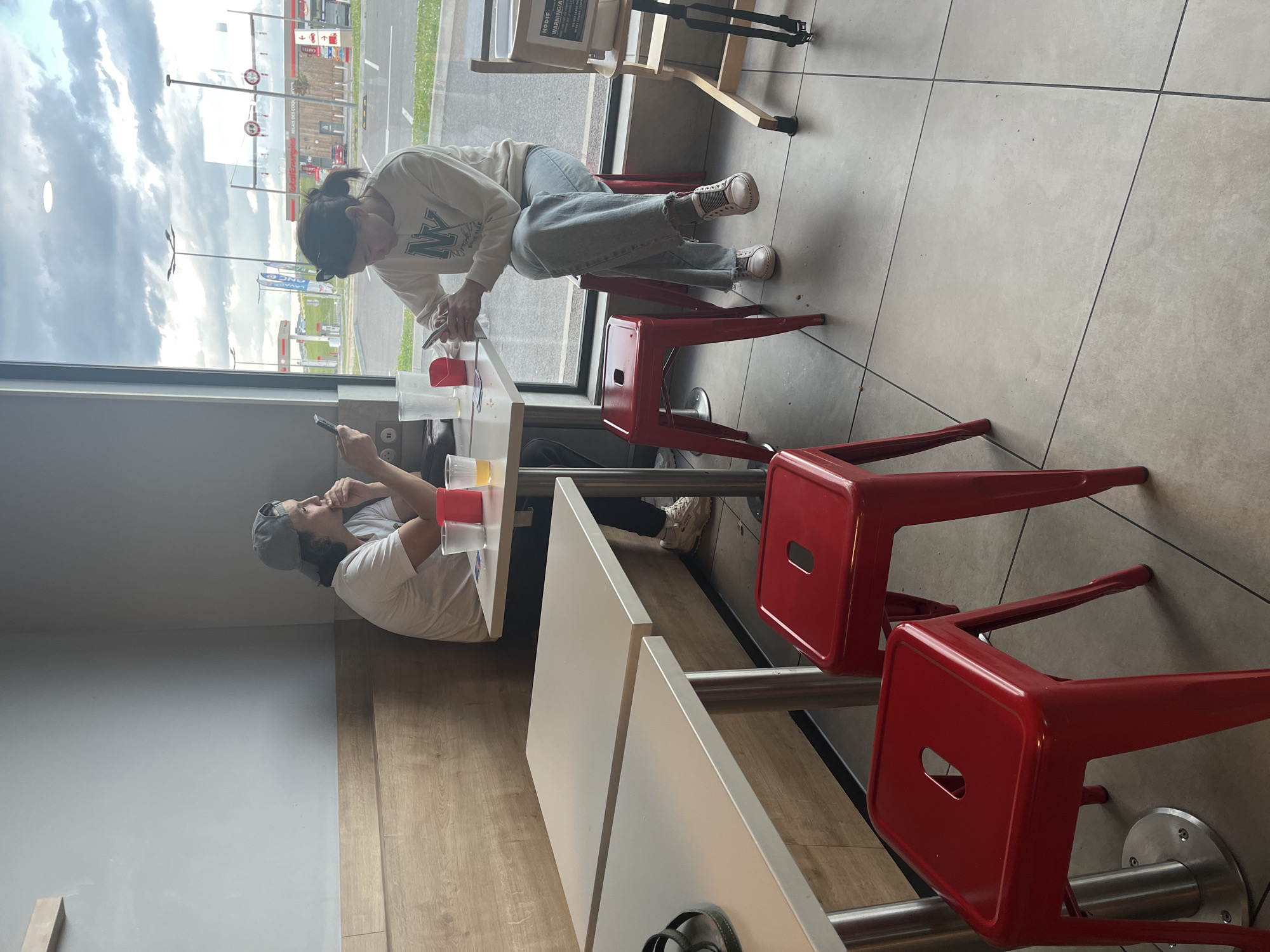 A photograph taken indoors shows two individuals seated at a white table in a brightly lit space. A woman, wearing a light-colored sweatshirt with "NY" written across it, light blue jeans, and white sneakers, is seated on a red stool and leans over the table, looking at her phone. A man, wearing a light-colored shirt and a baseball cap, sits across from her, also looking at his phone. The table holds several clear plastic cups, one with a yellow liquid. Additional red stools are positioned nearby.
Large windows reveal an exterior cityscape with a "Total Energies" gas station visible in the distance, along with a "ONCF" sign. The location is Andrézieux-Bouthéon, France, as indicated by the recognizable gas station and the ONCF sign. The interior has gray tiled flooring, a wooden accent wall, and white walls. A highchair is visible near the window.