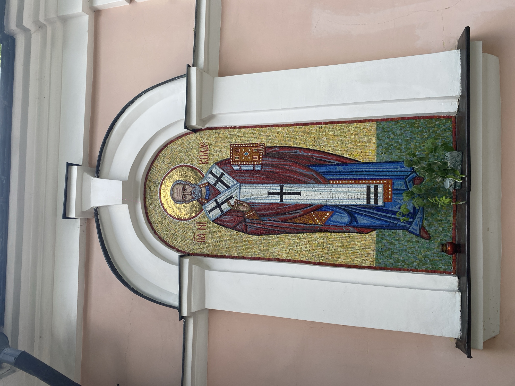 This image shows an exterior mosaic of Saint Nicholas on the facade of a building in Khimki, Russia. The mosaic depicts Saint Nicholas in traditional religious attire, including a red and blue robe adorned with crosses, and holding an open book. He has a white beard and is surrounded by a golden halo. The mosaic is set within an arched niche framed by white architectural elements and is mounted on a light pink building. A small, dark green plant is visible growing from a ledge below the mosaic, next to a red votive candle in a glass holder. The overall scene is one of religious art and architecture.