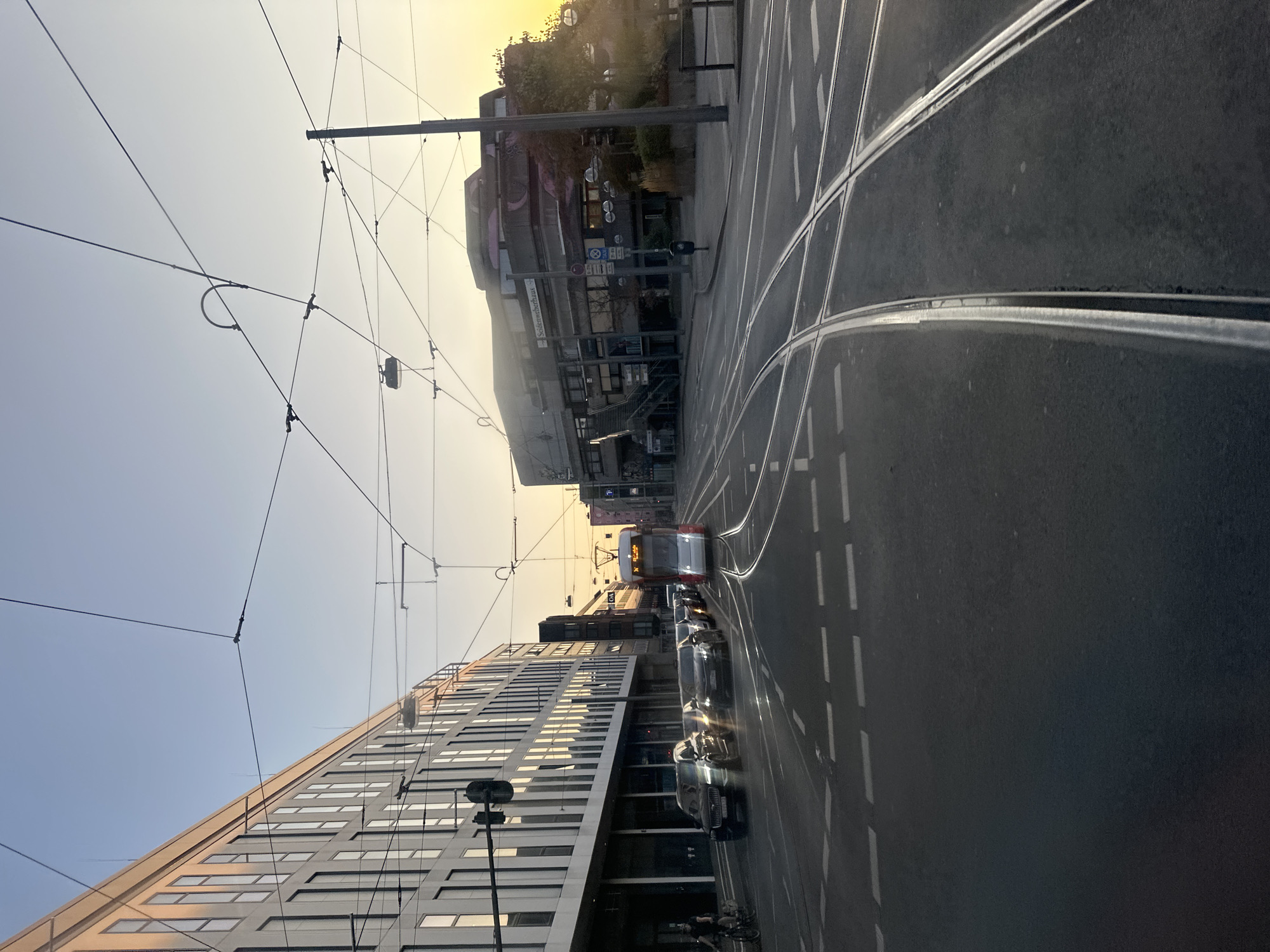 The image depicts a street intersection in Krefeld, Germany, likely during the golden hour due to the warm, low sunlight. A modern, multi-story building with numerous windows is visible on the left, reflecting the light. Across the street, a building with a unique, angular architectural style and graffiti on its facade stands. Tram tracks are prominent in the foreground, with a red and white tram approaching from the distance. Several cars are lined up on the street, indicating moderate traffic. Overhead, a complex network of tram wires crisscrosses the sky. A person on a bicycle is visible on the far left. The overall scene is urban, with elements of transportation, architecture, and daily city life under a soft, atmospheric light.