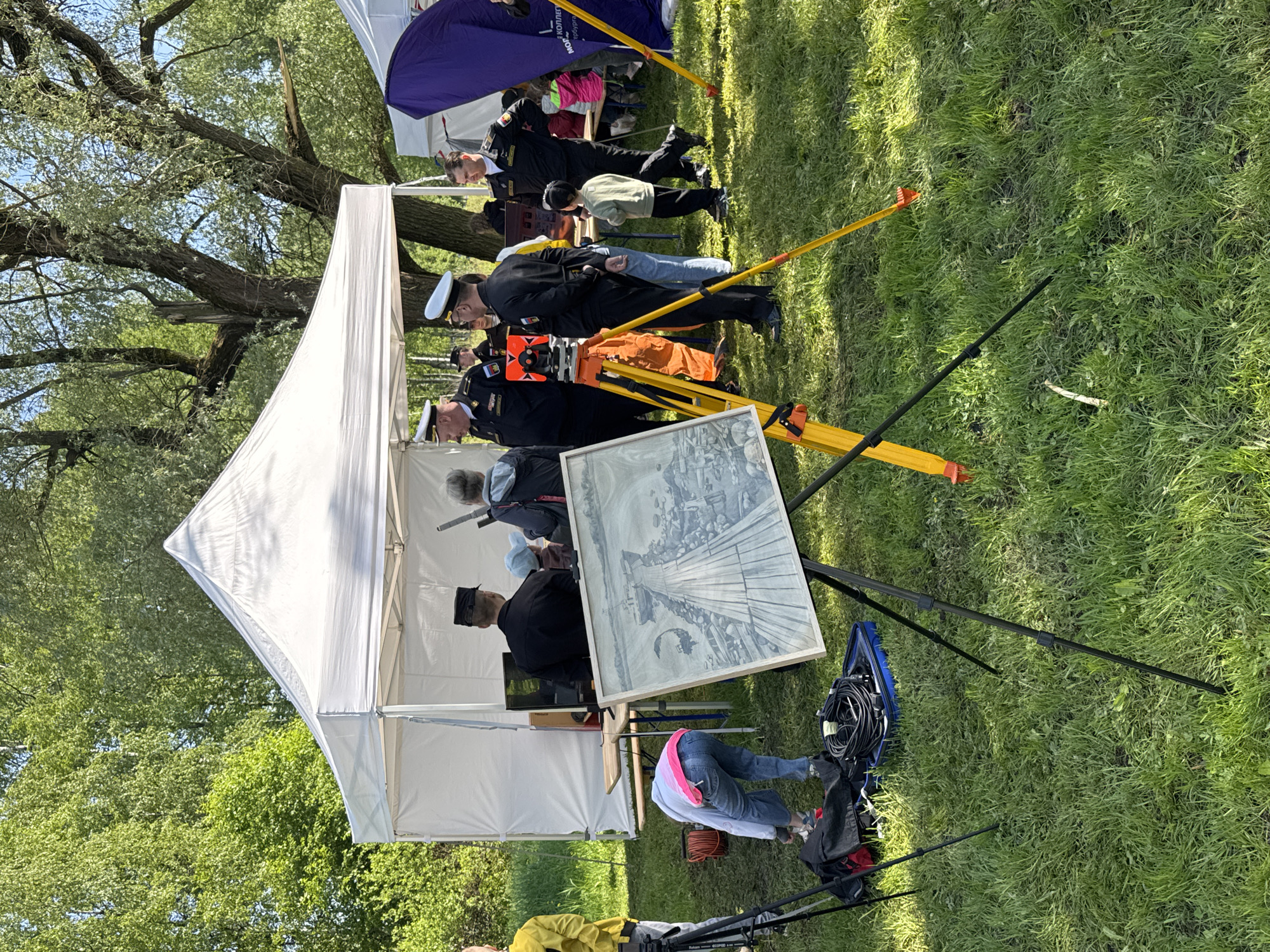 A ground-level, landscape-oriented, outdoor scene depicts a white canopy tent erected on green grass in Unknown, Unknown. To the right of the tent, a group of people wearing naval uniforms are positioned. A yellow tripod stands near the front of the tent. In front of the tent, there is a framed artwork depicting a black and white image of a pier or dock. Cables and equipment are piled near the bottom right corner of the frame. The grass is illuminated by sunlight. A tree is visible at the top left corner.