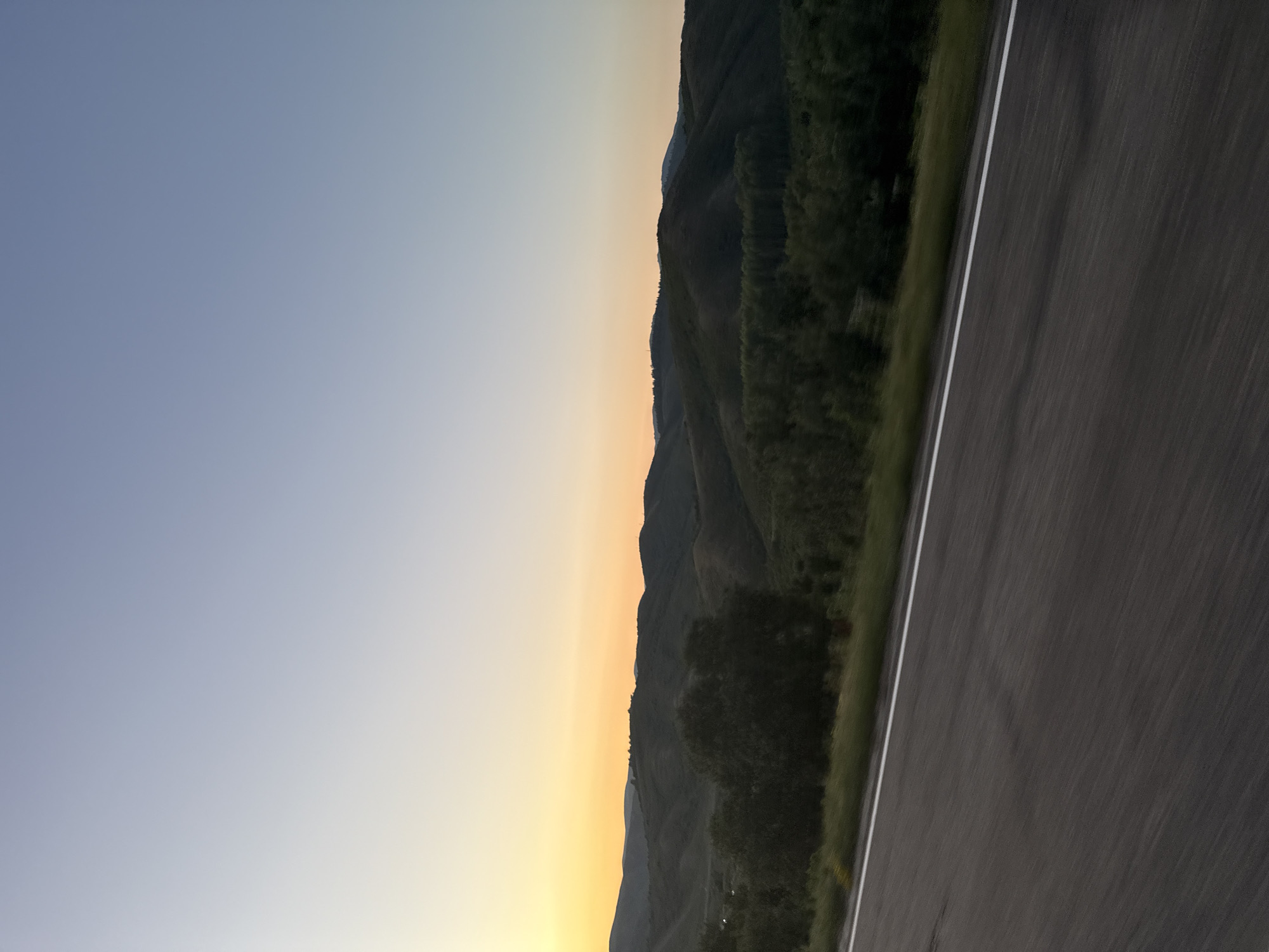 The image depicts a scenic landscape with rolling hills and a vast sky. In the foreground, a paved road with a white line marking the lane stretches into the distance. The road appears to be wet or reflective, suggesting recent rain or damp conditions. The road surface shows motion blur, indicating the photograph was taken from a moving vehicle.
Beyond the road, a strip of green grass and trees lines the roadside. The middle ground is dominated by a series of dark green, undulating hills covered in vegetation. The hills create a layered effect, with some appearing closer and more detailed than others, which fade into the distance. Scattered trees are visible on the slopes of these hills.
The background is a gradient of colors in the sky, transitioning from a pale yellow and orange hue near the horizon to a soft blue at the top of the image. This color gradient suggests the photograph was taken during either sunrise or sunset. The sky is clear, with no visible clouds.
The image does not contain any people, animals, or buildings. The overall environment is natural and undeveloped.
The location is identified as the city of Severnoe, Kazakhstan. However, the visual elements in the image (rolling hills, undeveloped landscape, paved road) do not provide specific landmarks or features that would definitively confirm this location without external information. The scene is characteristic of many rural or semi-rural mountainous or hilly regions.