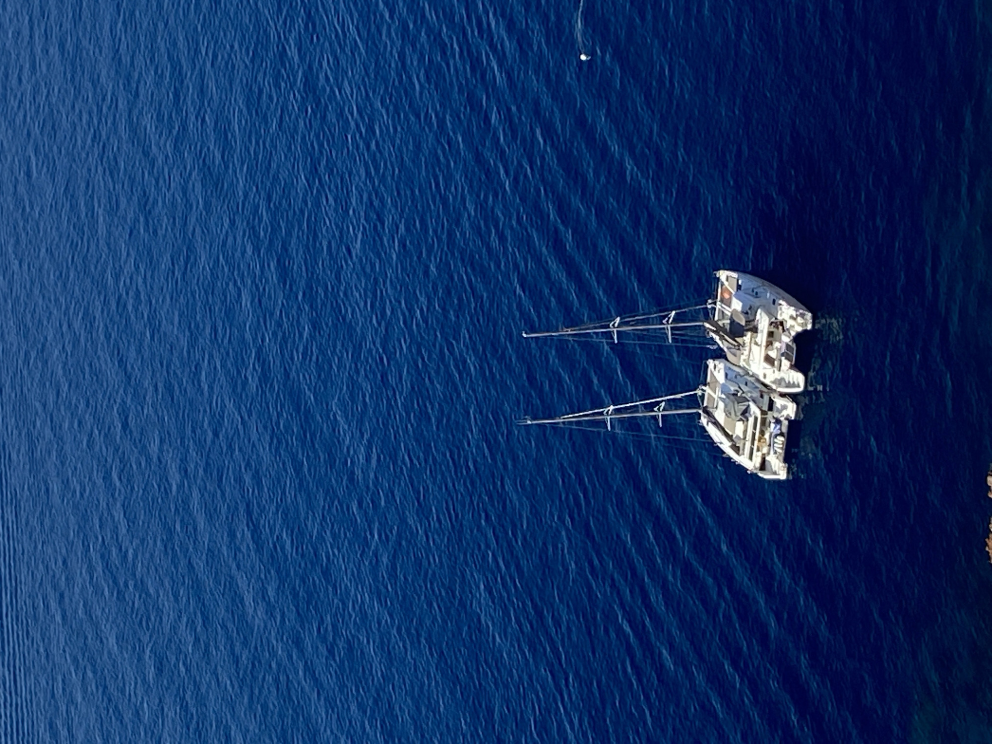 Two white catamarans are anchored in the deep blue Aegean Sea off the coast of Oia, Santorini, Greece. The water is calm with gentle ripples. The catamarans are positioned side-by-side, facing slightly to the left, with their masts erect. The foreground shows a hint of rocky coastline. The scene is a high-angle, aerial view.
