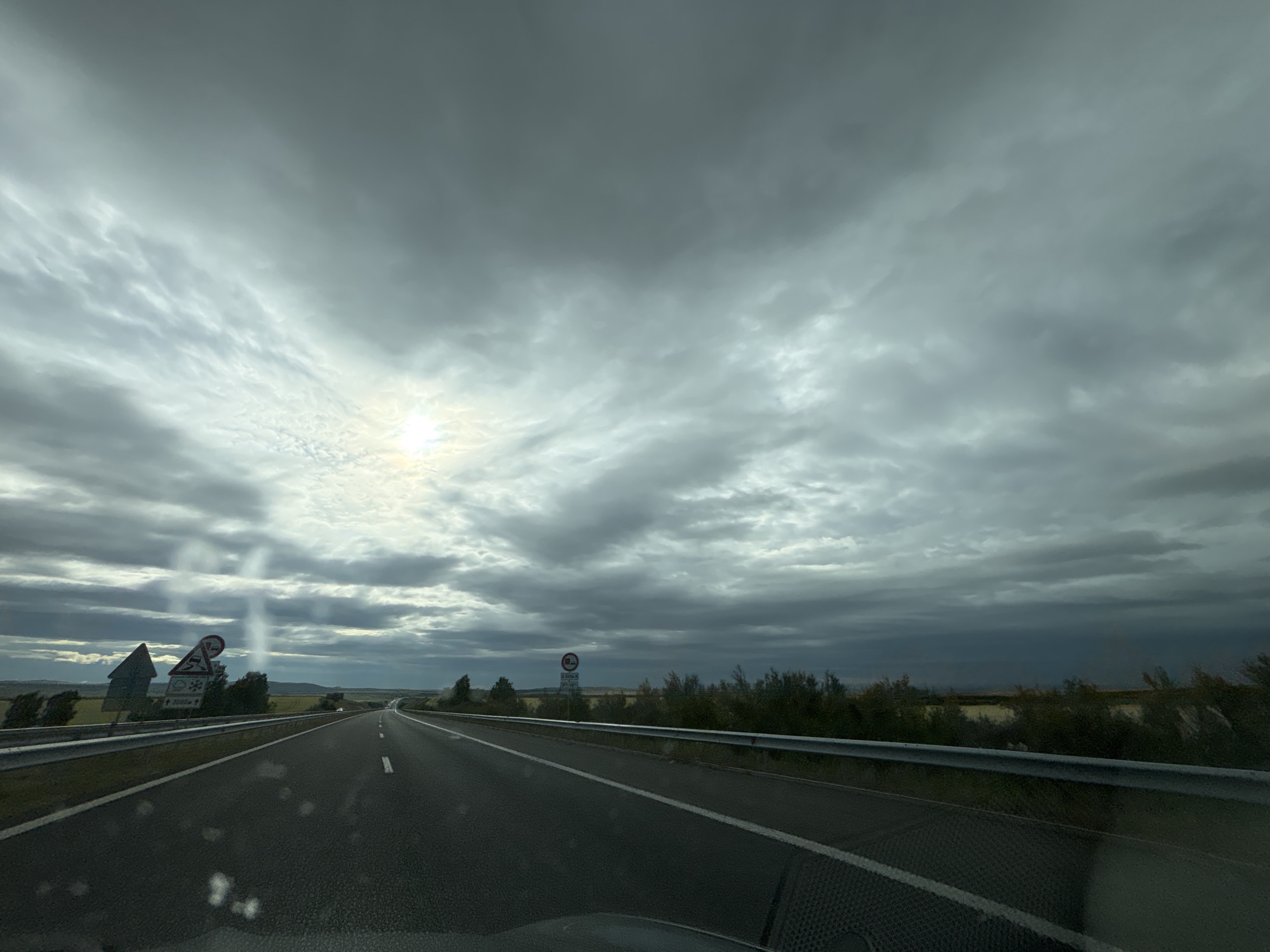 The image captures a view from inside a vehicle traveling on a highway, with a dramatic, cloudy sky overhead. The sun is partially visible, breaking through the clouds, casting a bright spot and creating some light rays. The highway stretches ahead, with multiple lanes and clear lane markings. To the left of the road, there are several road signs, including a directional sign with an arrow and a circular sign with a red border and a diagonal line, likely indicating a prohibition. Further down the road, on the right side, a metal guardrail runs alongside the highway, and a similar circular prohibition sign is visible. The landscape on either side of the highway appears to be somewhat barren, with low-lying vegetation and distant hills. The overall impression is one of a journey on a highway under an overcast and potentially stormy sky. There are no people or specific landmarks visible that would indicate the location in the city of Zheleznik, Bulgaria, or any other specific city. The image is a general highway scene with a focus on the weather conditions.