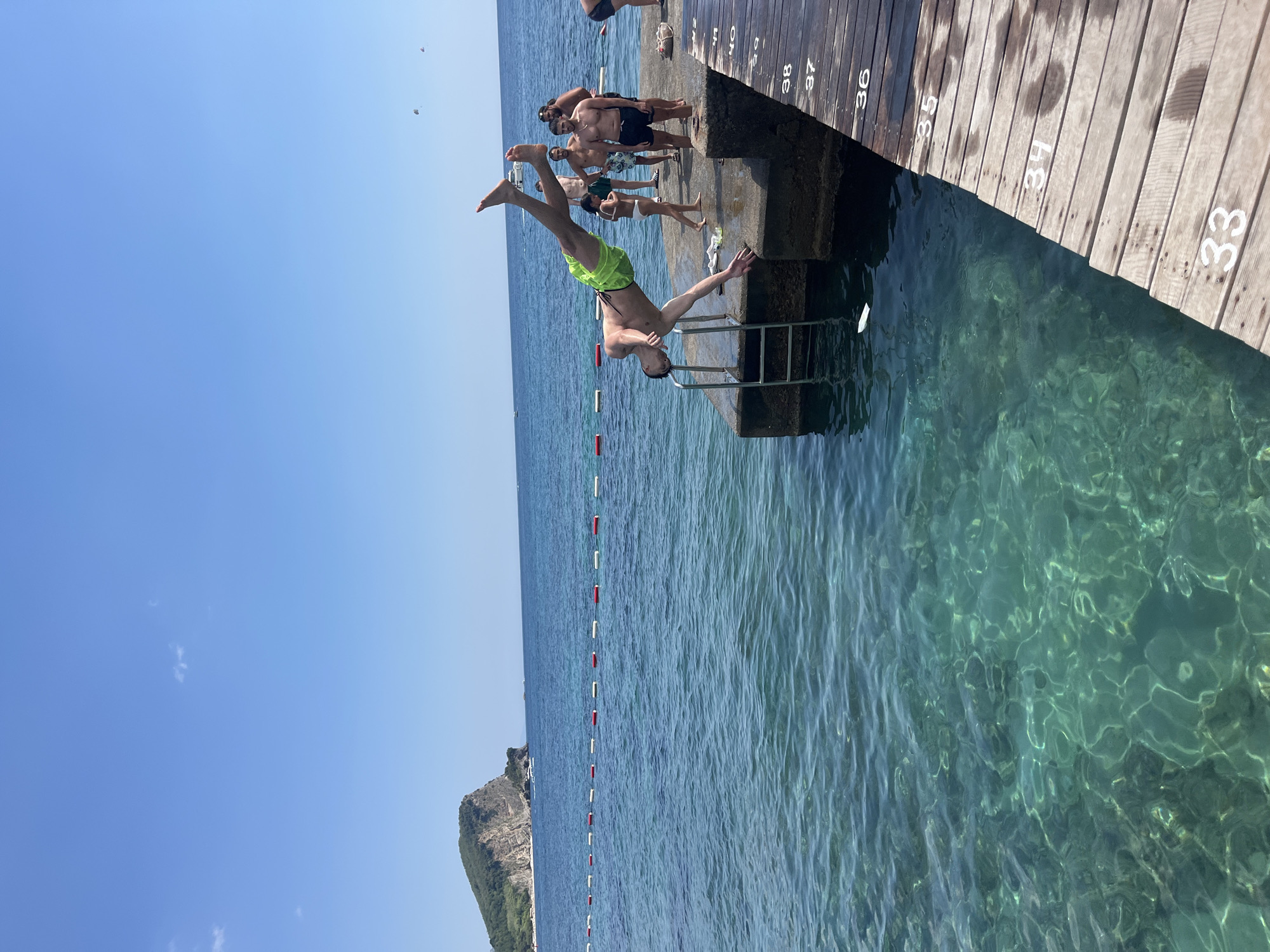 The image depicts a sunny day at a coastal location in Budva, Montenegro. The foreground is dominated by clear, turquoise water and a wooden pier. The pier has numbered sections, with visible numbers including 33, 34, 35, 36, 37, 38, and 40.
In the mid-ground, a young man in green shorts is captured mid-air, performing a dive from a concrete platform into the water. A metal ladder is attached to the side of the platform. Several other individuals are present on the platform and pier, observing the dive. These include a young girl in a white swimsuit, and several adult males in swimwear. One man is wearing black shorts, another is bare-chested with patterned shorts, and another is wearing a black cap and shorts.
The background reveals the Adriatic Sea, a rocky, tree-covered island with a small structure on it, and a clear blue sky. There are also visible buoys in the water, creating a boundary. In the far distance, there appear to be paragliders in the sky and small boats on the water. The overall scene is one of recreational activity at a seaside resort.