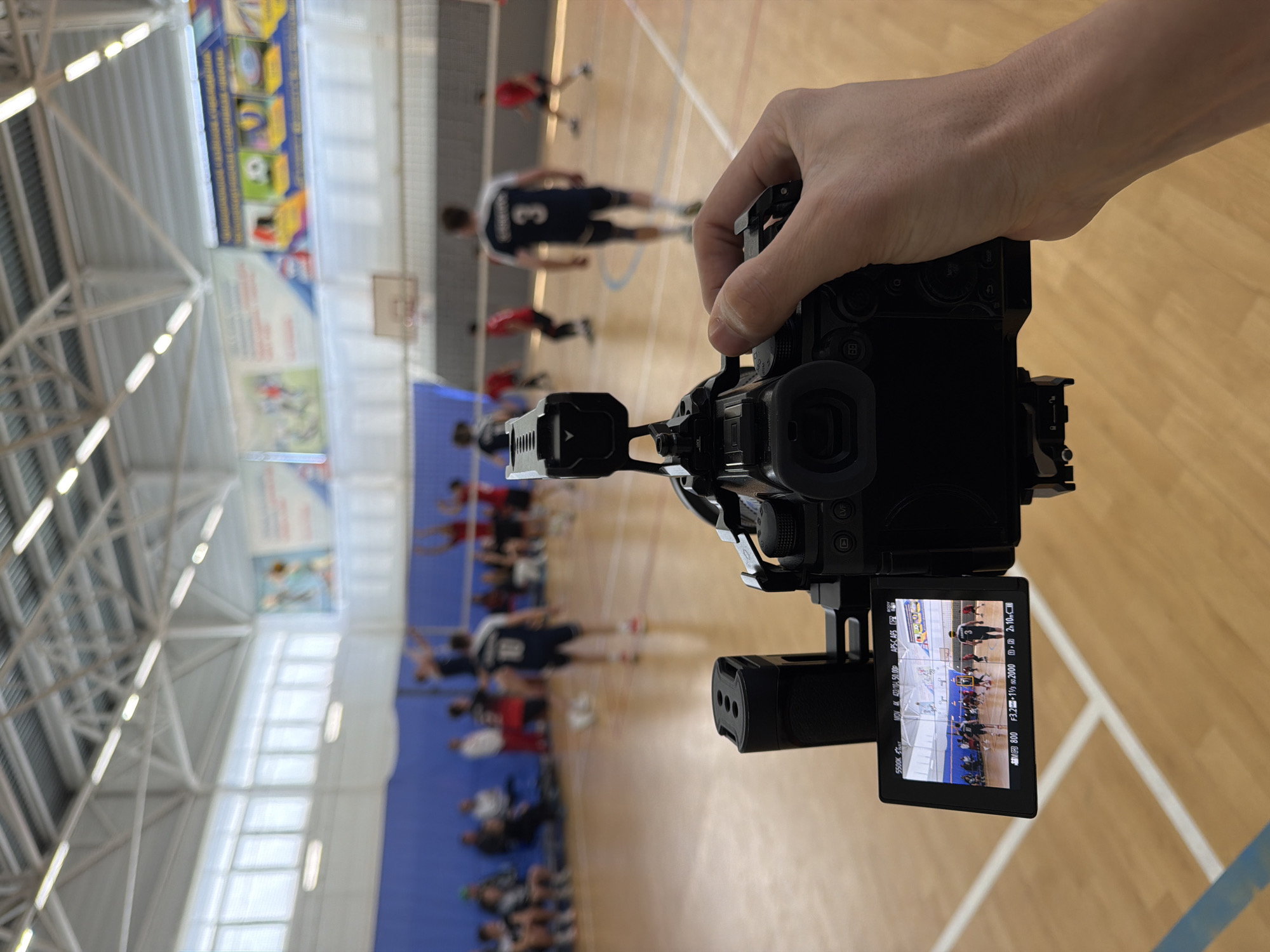 The image shows a person holding a camera in a gymnasium in Bolshaya Murta, Russia. The camera's display screen shows a basketball game being filmed. Players in jerseys are on the court, with spectators seated in the background. The gymnasium has a light-colored wooden floor with lines, blue walls, and a white ceiling structure with overhead lights. Banners are displayed on the walls.