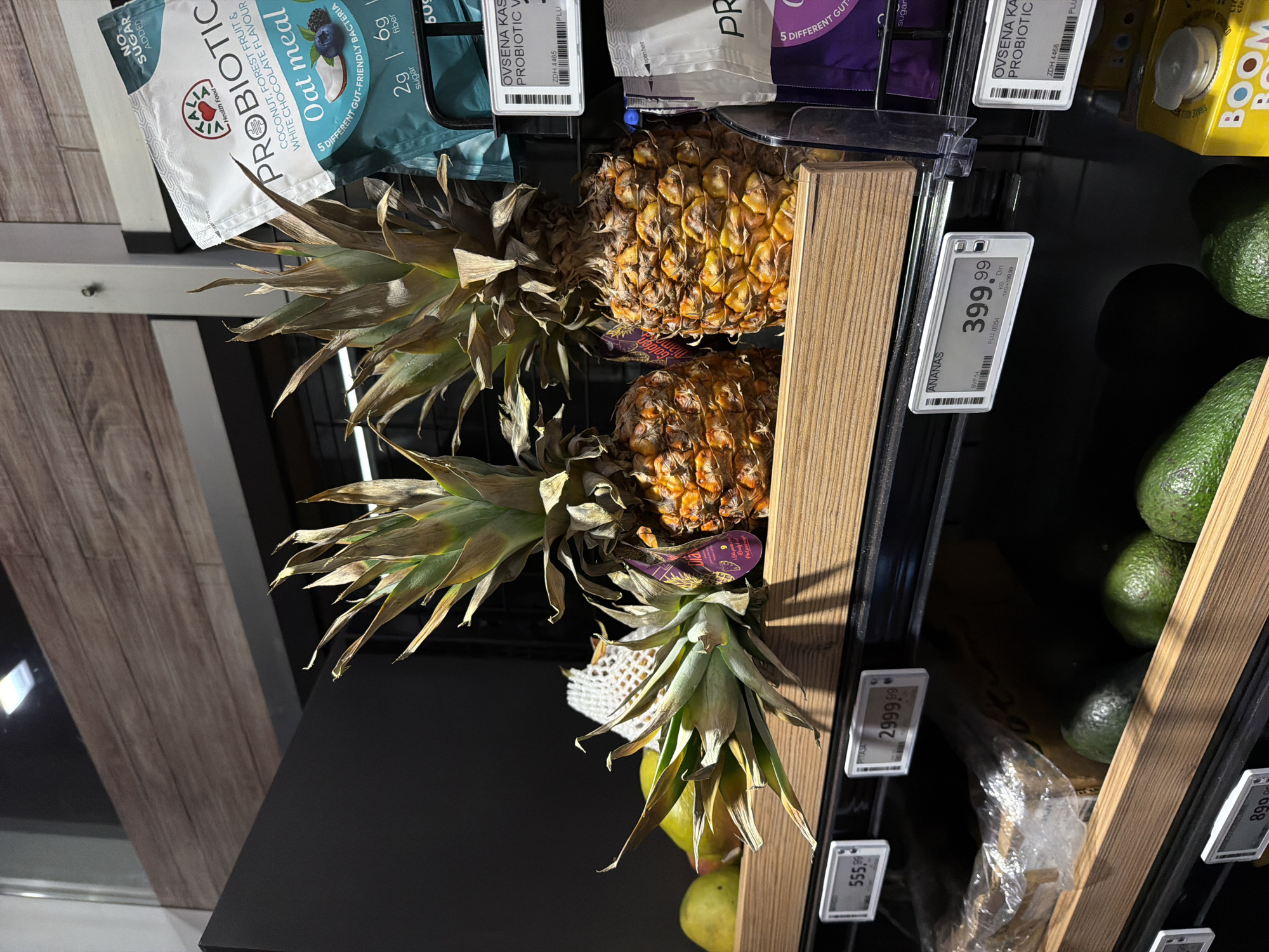 The image shows a shelf in a store. The products on the shelf include pineapples, avocados, probiotic oatmeal, and "Boom" juice. There are digital price tags on the shelf that say "Ananas 399.99", "PITAJAN 555.99" and "2999.99". This image was likely taken in a store in Vracar Urban Municipality, Serbia because the price tags are in Serbian currency (RSD).
