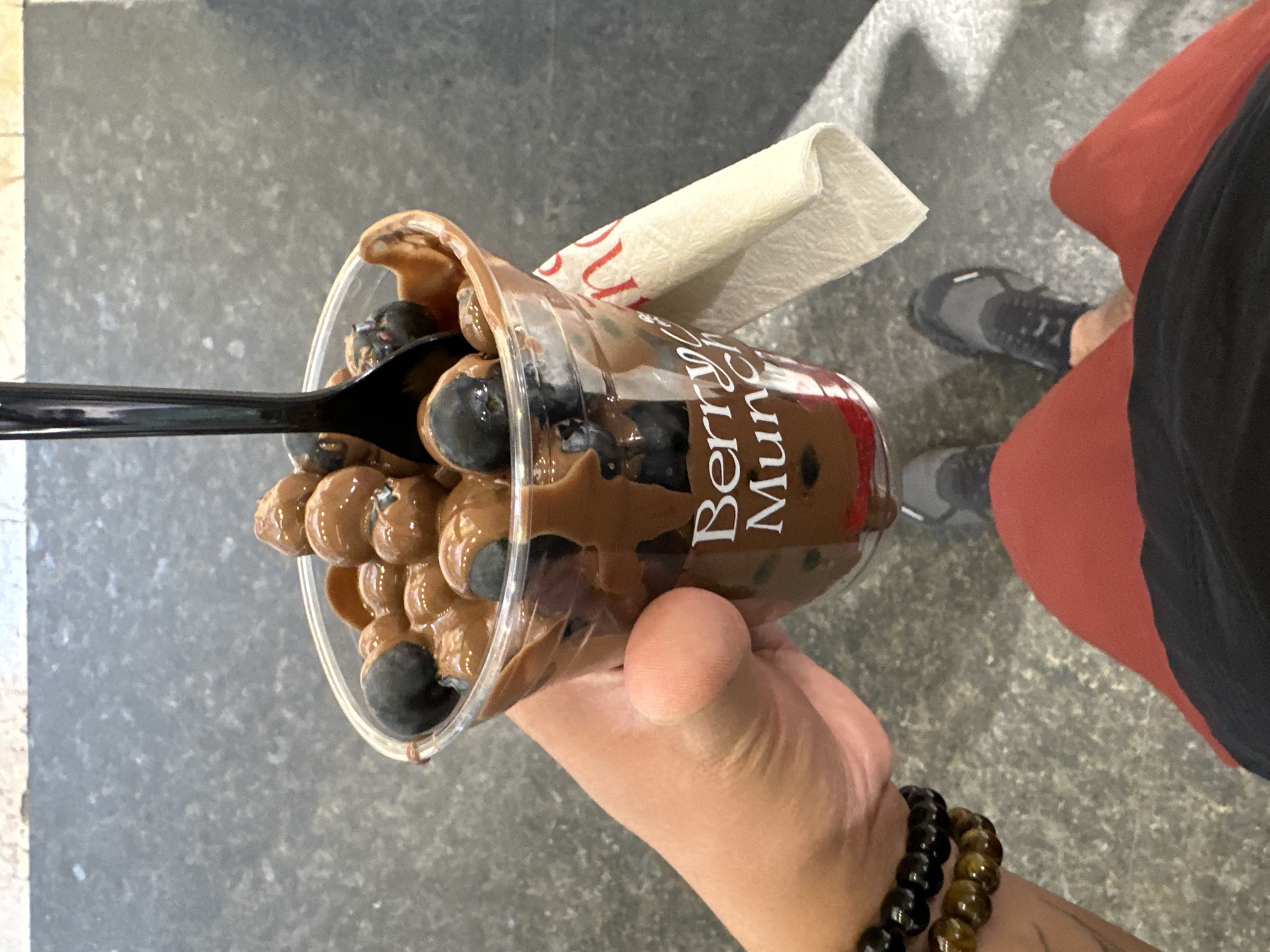 The image presents a close-up, downward perspective of a person's right hand holding a clear plastic cup. The cup contains blueberries generously coated in a viscous brown chocolate-like sauce, with a black plastic spoon inserted into the contents. White text reading "Berry Munch" is visible on the side of the cup. A folded white paper napkin, partially obscuring red lettering, is tucked into the cup's side. The individual's right wrist wears two beaded bracelets, one black and one brown, with a faint tattoo visible on the forearm. Below the cup, the person's lower body is visible, clad in red shorts and black and gray athletic shoes. The flooring is a speckled dark gray, consistent with polished concrete or commercial tiling. A light-colored tiled baseboard is discernible along the top edge of the frame. This scene depicts an indoor commercial setting, typical of a food vendor kiosk or food court within a shopping mall in Puebla, Mexico, specifically at a location where Berry Munch operates, such as the Angelópolis Lifestyle Center.