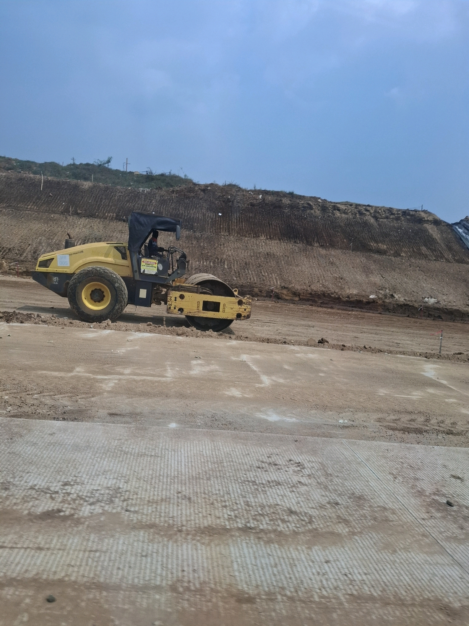 An image depicts a bright yellow road roller parked on a dirt surface with a large, excavated hillside in the background. The sky is clear and blue. The road roller is positioned slightly to the left of the frame, with its large rear drum and front wheel visible. A black canopy is extended over the operator's cabin, where a person appears to be seated. The hillside behind the roller shows visible layers of disturbed earth, suggesting ongoing construction or excavation. The foreground is a rough, unpaved ground surface, with a section of textured, possibly cured concrete in the lower right corner. Red and white markers are sporadically placed along the base of the hillside and in the mid-ground. The scene appears to be a construction site in Karangtengah, Indonesia.
