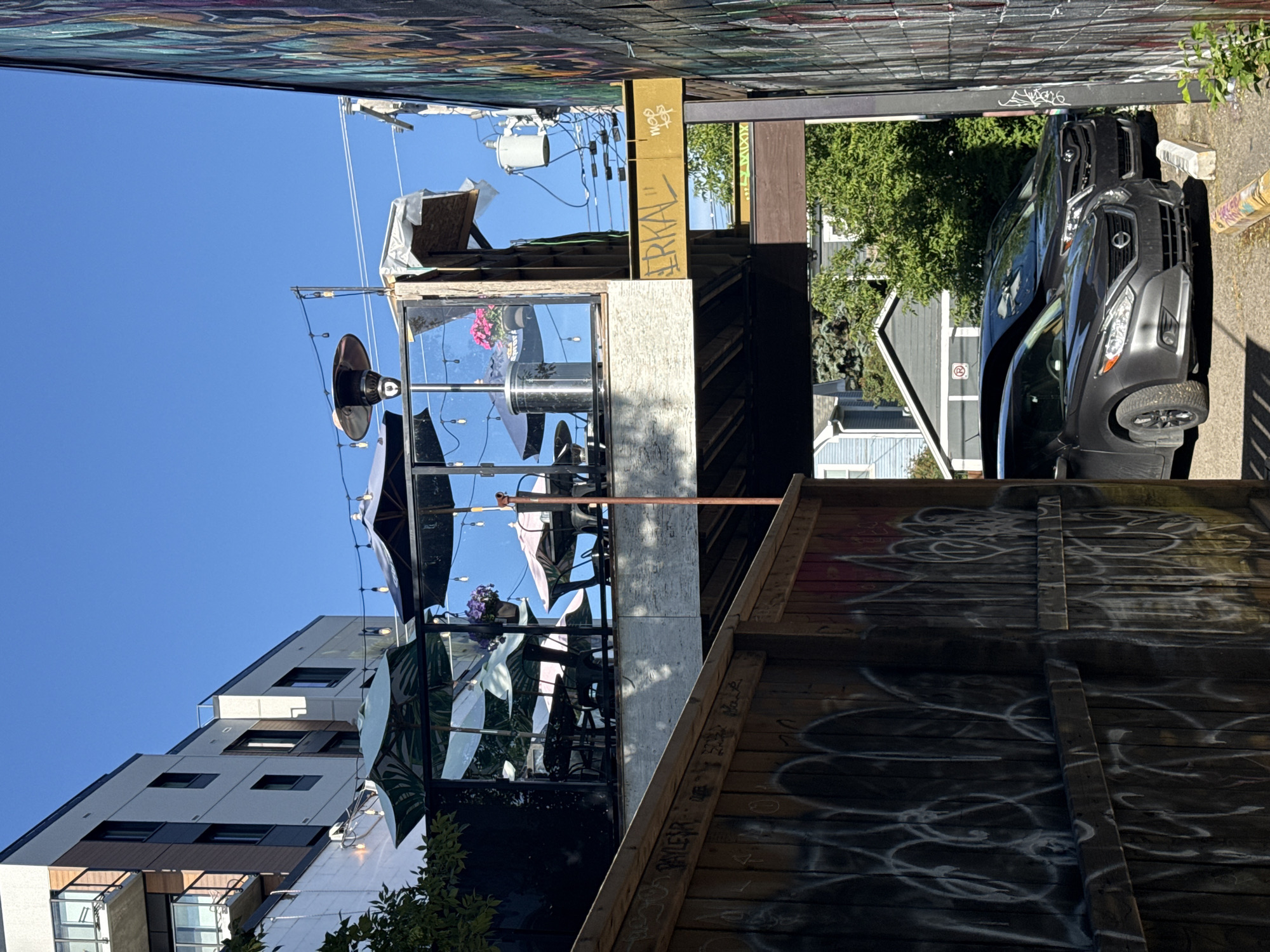 The image shows an outdoor patio area on an elevated platform, likely part of a restaurant or bar, with tables and umbrellas visible. A patio heater is mounted on the platform. Below the patio, there is a narrow alleyway where two cars are parked. One car, a dark gray sedan, is in the foreground, and a dark gray minivan is parked behind it. The alley is lined with graffiti-covered wooden fences on the left and a graffiti-covered wall on the right. The background shows buildings and trees, and the sky is clear and blue. The sign on the yellow beam of the structure above the alley reads "ERKAL" with "MOP TOP" written below it, possibly indicating street art or a business name. Based on the distinctive architectural style of the building on the left and the presence of similar street art in the area, this scene is likely located in the Beltline neighborhood of Calgary, Canada, specifically near the intersection of 17th Avenue SW and 1st Street SW.