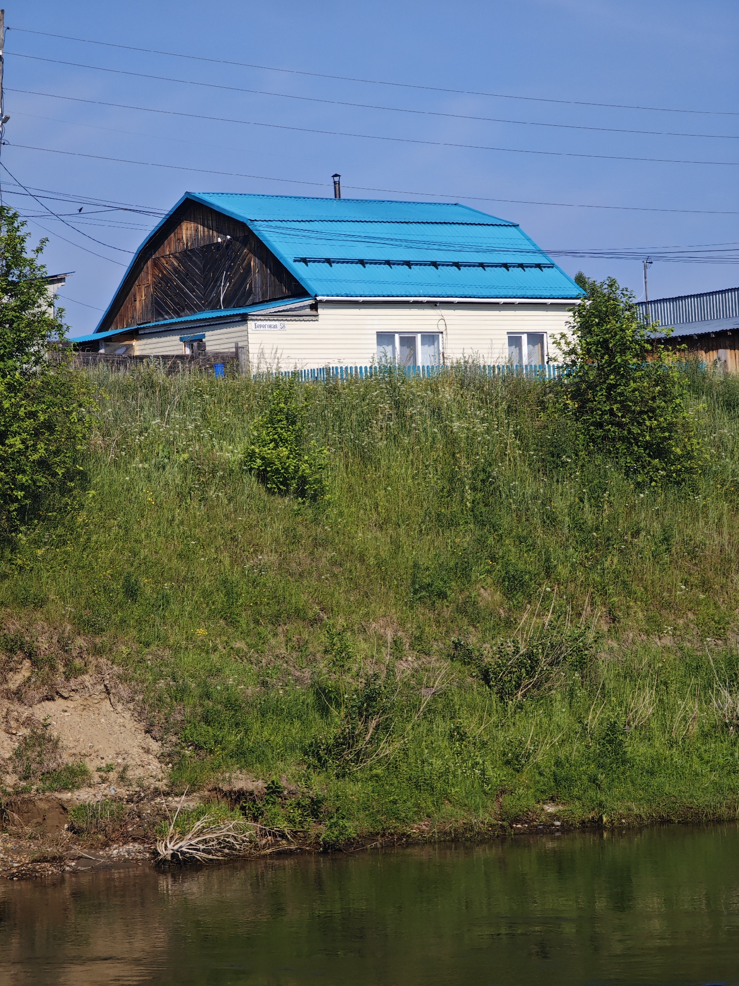 The image captures a view of a house with a bright blue, corrugated metal roof, situated on a grassy embankment overlooking a body of water. The house is painted cream with white trim around two windows and a blue picket fence runs in front of it. A street sign reading "Береговая 58" (Beregovaya 58) is visible on the side of the house, indicating its address. The embankment is covered in lush green grass and wildflowers, with a sandy, eroded slope on the left leading down to the water's edge. The water's surface is dark and reflective. In the background, power lines stretch across a clear blue sky. To the right of the main house, another building with a blue-striped roof is partially visible. The scene is consistent with a residential area in a rural or suburban setting, possibly in or near Galanino, Russia, given the street sign. There are no people or animals visible in the image.