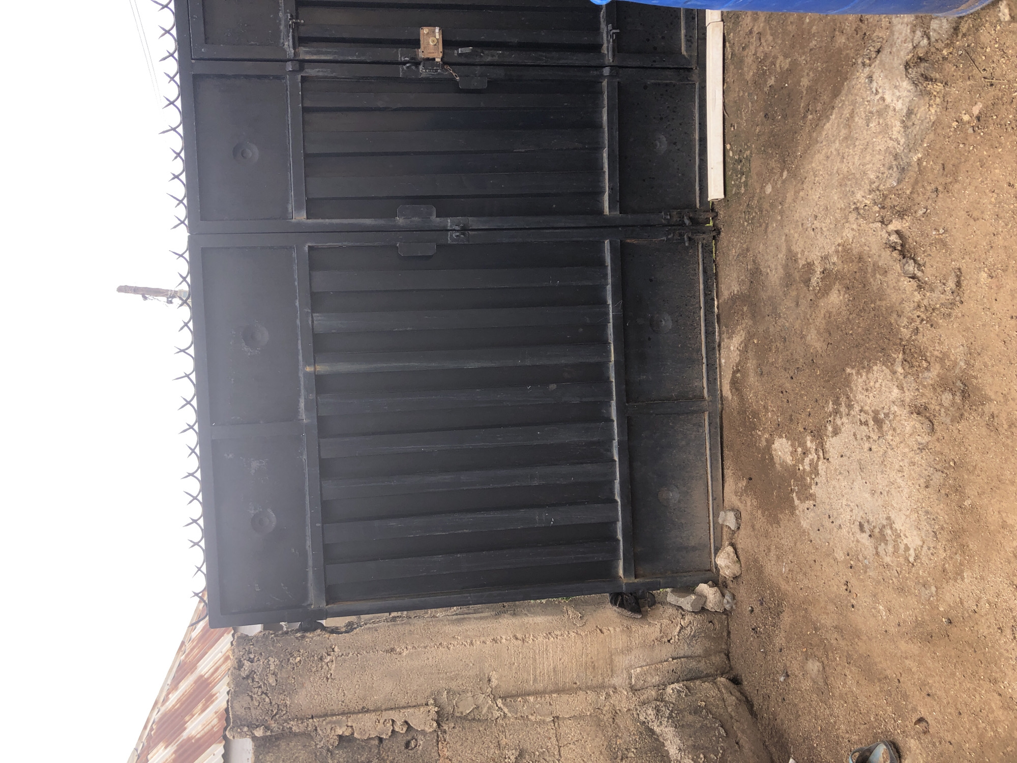 The image depicts a secured entryway to a property in Kabong, Nigeria, under an overcast sky. A dark grey or black metal gate, composed of two main sections, dominates the center. The left gate section features vertical corrugated panels below a flat rectangular panel with two circular indentations. The narrower right section, also with vertical corrugated panels and a flat upper panel, is secured with a visible padlock and chain. Above the entire gate structure, a line of razor wire is installed for security.
To the left of the gate, a rough-textured, weathered concrete or mud-brick wall forms part of the property boundary. Partially visible above this wall and the left side of the gate is a rusted, corrugated metal roof.
The unpaved ground in the foreground consists of reddish-brown dirt, compacted soil, and gravel, with scattered small stones, debris, and dark specks resembling animal droppings. Patches of moisture or darker earth are visible on the ground. A light blue open-toed sandal is partially visible in the bottom-left corner, and a blue plastic barrel or container is partially visible in the bottom-right corner. A white strip, possibly a discarded PVC pipe, lies on the ground directly in front of the right gate section. No individuals or active animals are present in the frame.