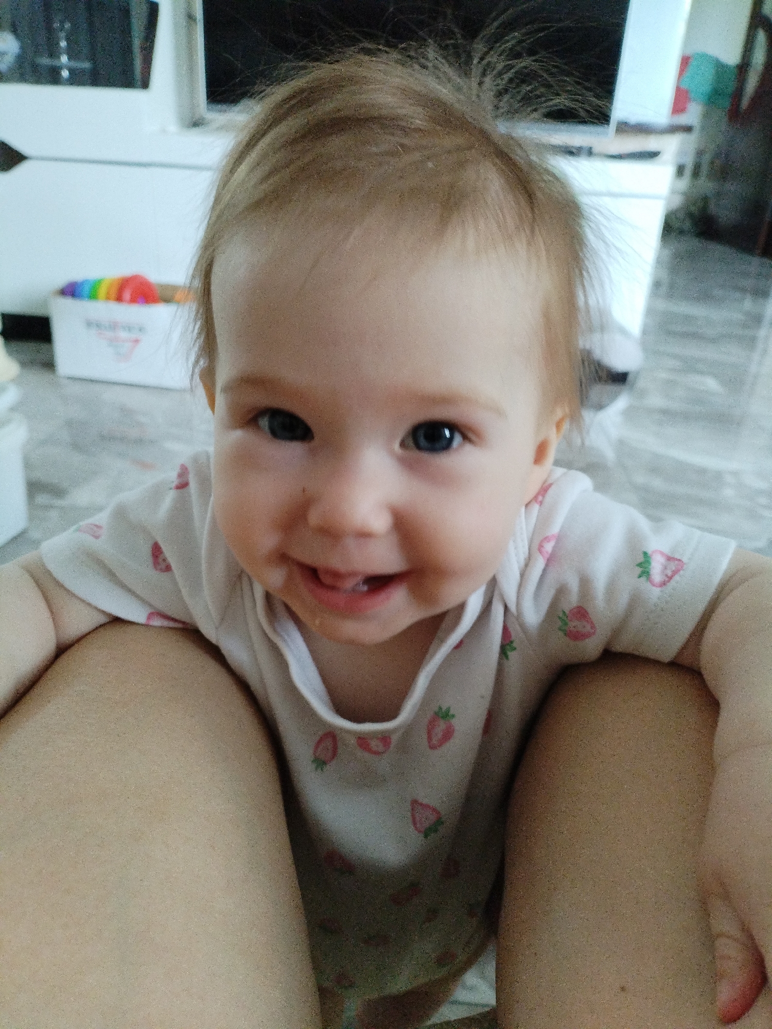 The image shows a close-up of a baby girl looking directly at the camera. She has fair skin, blonde hair, and blue eyes. She is wearing a white onesie with a pattern of pink strawberries. Her mouth is slightly open, revealing her tongue and teeth, and she appears to be smiling or laughing. The background is blurred, but it seems to be an indoor setting with some furniture and a toy in a box with a rainbow pattern. The lighting suggests it is daytime. The image appears to be a casual snapshot, possibly taken by a parent or caregiver. The baby is positioned in a way that suggests she is being held, with adult legs visible on either side of her. There is no visible text in the image.