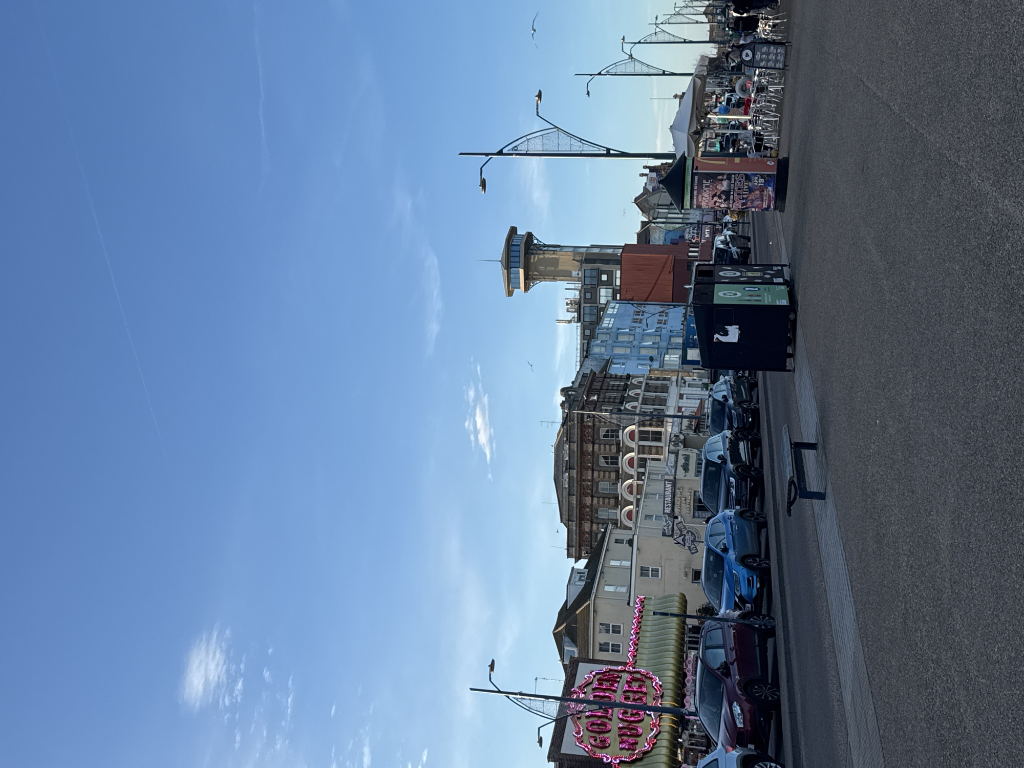 The image depicts a coastal scene in Gorleston-on-Sea, United Kingdom. The sky is clear blue with a few scattered clouds and contrails. Along the right edge of the image, there is a paved road or walkway. Cars are parked along the street. Buildings of varying heights and architectural styles line the street, including a tall tower structure with a viewing platform. Streetlights with distinctive arched supports are visible. A brightly lit sign for "Coffee Corner" stands out near the bottom of the image. There are some people on the sidewalk but are small and cannot be recognized. There are a few birds in the sky. A black trash container is also visible.