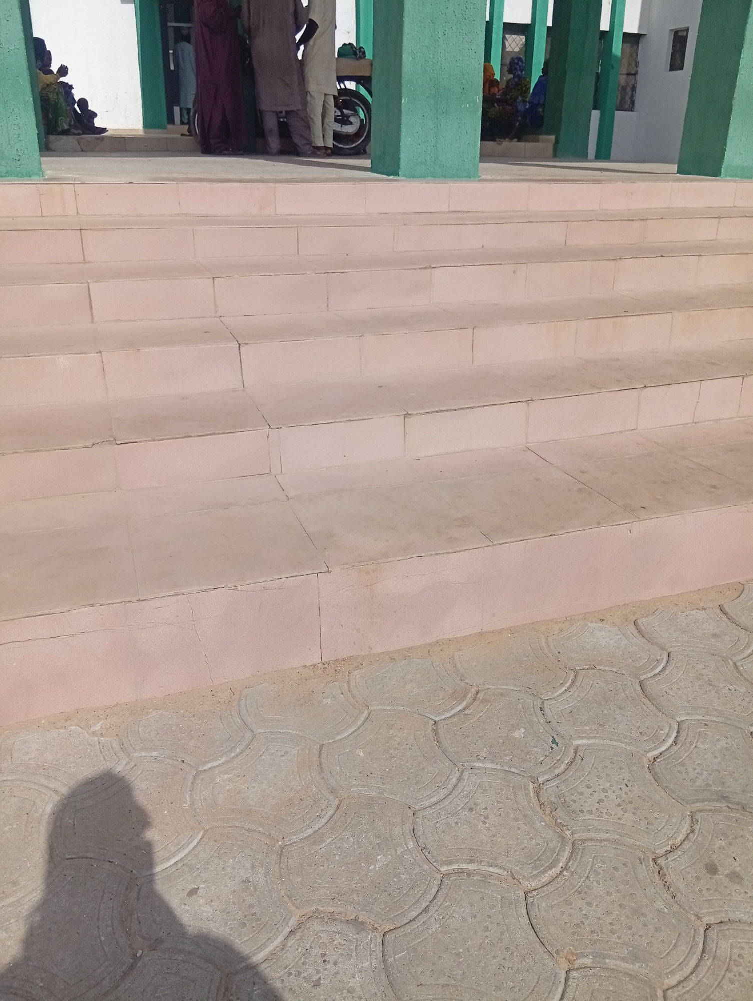 The image captures an outdoor scene featuring a flight of light pink/beige tiled stairs leading up to a building entrance. The building is characterized by prominent green-painted pillars supporting an overhang. The ground area in front of the stairs is paved with gray, interlocking, scalloped-edge concrete tiles.
Several people are gathered on the landing at the top of the stairs, partially visible behind the green pillars. They appear to be dressed in traditional long robes, with colors including maroon, brown, and light beige. Some individuals are standing, and one figure on the left appears to be seated or crouching, possibly with a child nearby. A motorcycle or scooter with a green component on its rear is partially visible among the people.
The activity taking place seems to be a gathering or waiting at the entrance of the building. The bright sunlight and a distinct, long shadow of a person, likely the photographer, cast on the tiled ground in the foreground, indicate it is daytime, possibly morning or late afternoon. There is no visible text in the image. The setting and attire are consistent with the provided location context of Birnin Kudu, Nigeria.