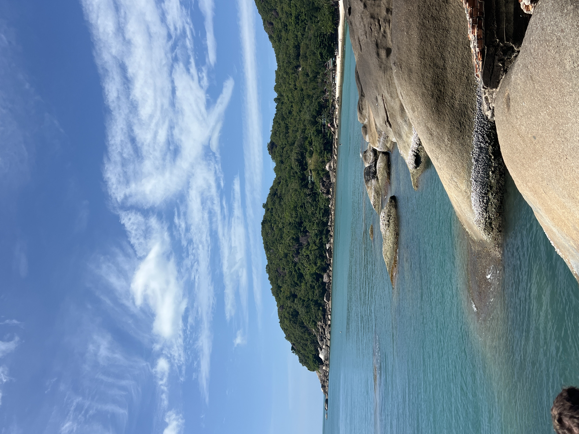 This is a photograph taken from the coast of Koh Samui District, Thailand, likely during daylight hours. The image showcases a picturesque tropical scene with a clear blue sky dotted with wispy white clouds. The foreground is dominated by large, smooth, grey boulders that extend into the turquoise water. The water is calm with some visible ripples around the rocks.
In the mid-ground, the coastline curves, revealing a sandy beach lined with palm trees and some buildings, possibly resorts or restaurants. Lush green, tree-covered hills form the backdrop, rising steeply from the shoreline. The sea itself is a vibrant shade of turquoise, indicating shallow, clear waters. There are no people clearly visible in the immediate foreground, but faint figures can be seen on the distant beach. The overall impression is one of a serene and beautiful natural landscape.