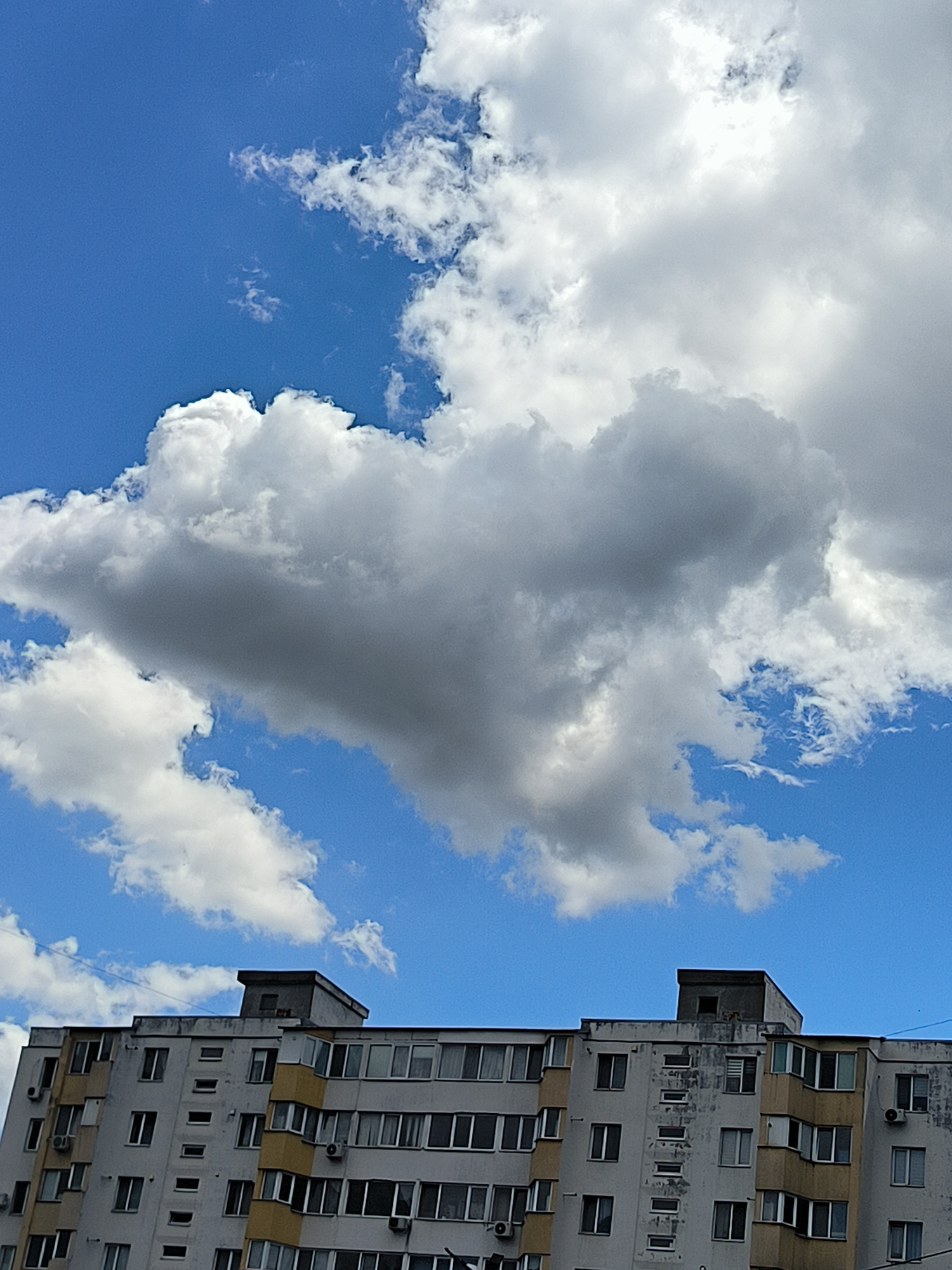 A low-angle shot captures a section of a multi-story apartment building against a bright blue sky filled with dramatic, puffy clouds. The building, a typical Soviet-era structure, is light-colored with yellow accents around some of the bay windows. Numerous rectangular windows, some with air conditioning units beneath them, punctuate the facade. The uppermost section of the building features a darker, more utilitarian structure with a small window. The clouds dominate the upper two-thirds of the frame, with a large, dark gray cloud formation casting a shadow, creating a stark contrast with the vibrant blue sky and bright white clouds elsewhere. The scene appears to be a residential area in Tiraspol, Moldova, likely captured on a partly cloudy day. There are no people or animals visible in the image.