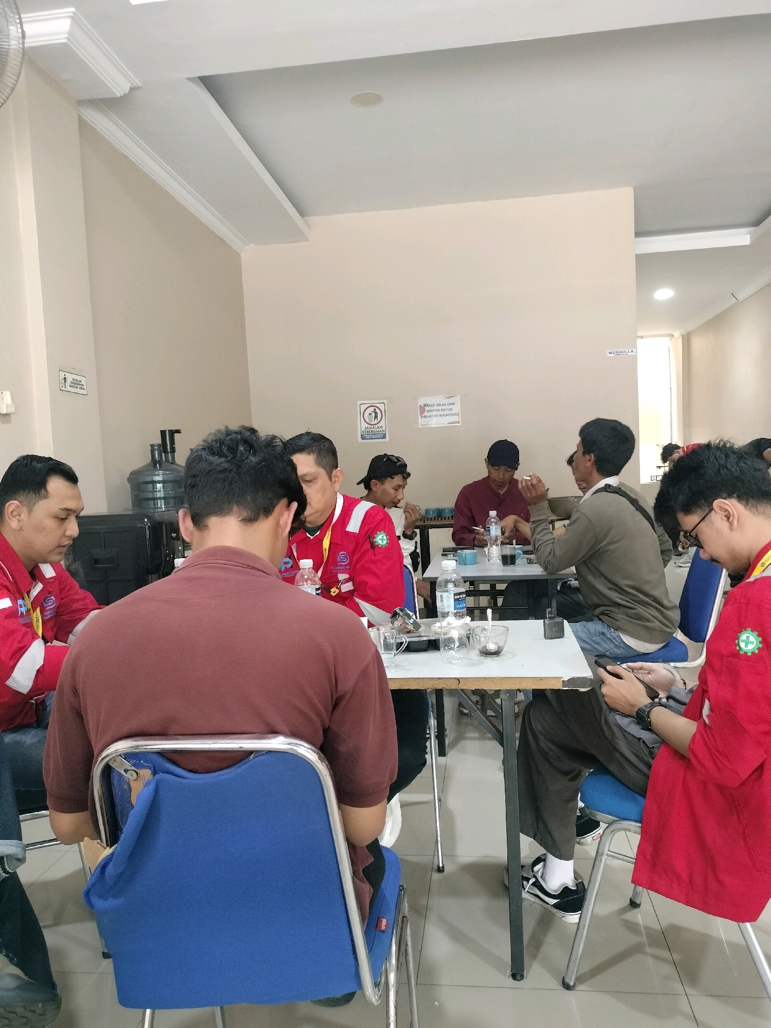 The image depicts an indoor scene, likely a break room or common area, with several individuals gathered around tables. The environment appears to be a commercial or industrial setting, suggested by the presence of water dispensers, white tables, and chairs that resemble those found in such facilities.
In the foreground, a person wearing a maroon polo shirt is seated on a blue chair, facing away from the camera. To their left, a man in a red uniform with white stripes on the sleeves is seated at a table, appearing to be in conversation or engaged with something on the table. Further to the left, another man in a similar red uniform is also seated at a table, looking downwards.
In the middle ground, a group of men are seated around tables. One man in a gray sweater and blue jeans is holding a phone. Another man in a dark maroon shirt and a black cap is seated and appears to be smoking. A man in a red uniform is visible on the right side of the image, also wearing a blue chair.
The walls are a neutral, light color, and the ceiling has modern lighting fixtures. There are signs on the wall, one with text in Indonesian and an icon suggesting cleanliness. Another sign has text that appears to be "MUSHOLLA," indicating a prayer room. The floor is tiled in a light color.
While the image shows people in what could be a communal setting, specific details about their interactions or the nature of their gathering are not fully discernible. The presence of uniforms suggests they may be employees of a company or members of an organization. The setting itself does not provide definitive clues to a precise location within Batam, Indonesia, beyond being an indoor common area.