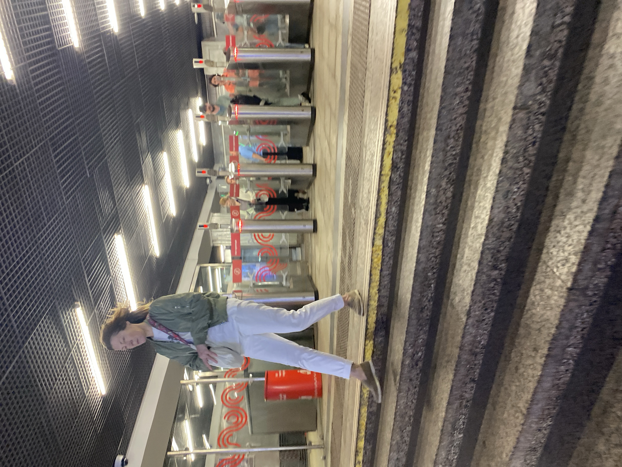 In Khimki, Russia, a woman walks down a set of stairs in a metro station. She is wearing a green bomber jacket, white pants, and beige slip-on shoes. She carries a white crossbody bag. Behind her, several turnstiles with red and white lights are visible, with other people passing through them. The station features a dark, grated ceiling with fluorescent lighting. The stairs are made of stone and have yellow safety markings on the edges.