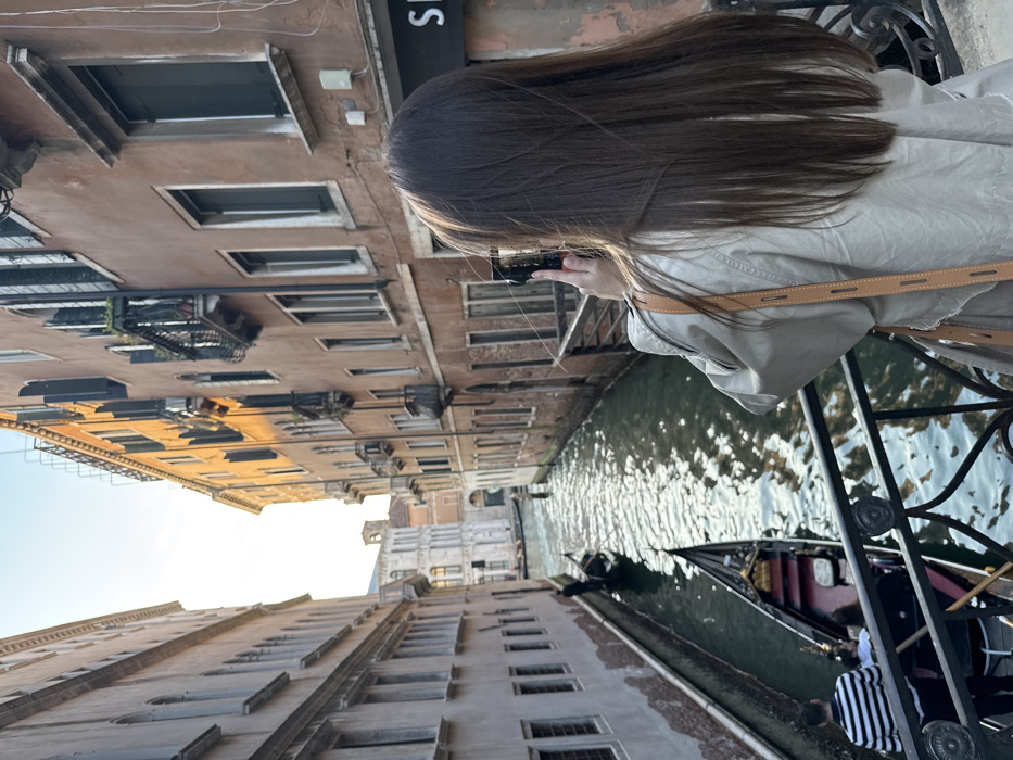 A forensic description of the scene in Venice, Italy, reveals a female individual standing on a bridge, facing away from the viewer, actively photographing a canal with a dark-colored smartphone. Her long, straight, dark brown hair falls over the shoulders of a light-toned trench coat, accented by a light brown leather strap. The bridge features ornate black metal railings with circular decorative elements.
Below, a narrow canal reflects sunlight on its green-blue surface. Two black gondolas are present: one closer to the bridge carries at least one passenger (wearing a black and white striped shirt, seen from behind) and a gondolier (partially visible), while a second gondola further down the canal is also occupied by a gondolier.
The canal is flanked by multi-story buildings. On the left, a building with a light stucco facade displays numerous rectangular windows. On the right, a building with a reddish-brown facade features dark window shutters, small balconies with black railings, and a partially visible ground-floor sign reading 'S H'. Distant buildings, including a prominent light-colored bell tower, are visible against a bright, clear sky at the end of the canal.