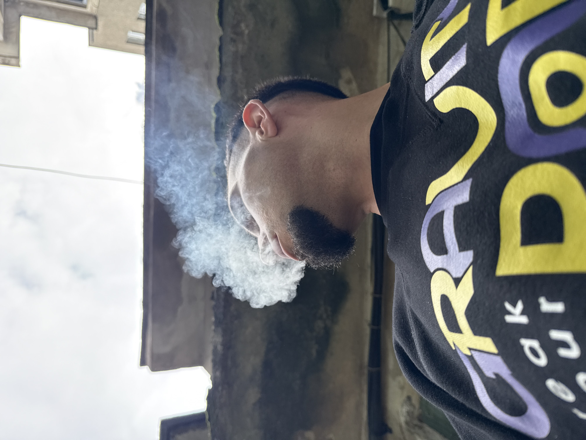 A low-angle, close-up photograph depicts a man exhaling smoke. The man, seen from the side, has a closely cropped haircut and a dark mustache. He is wearing a black shirt with yellow and purple lettering. The background includes a weathered concrete wall and a building facade with a cloudy sky visible above. There is not enough information to discern the location in the city of Shumen, Bulgaria.