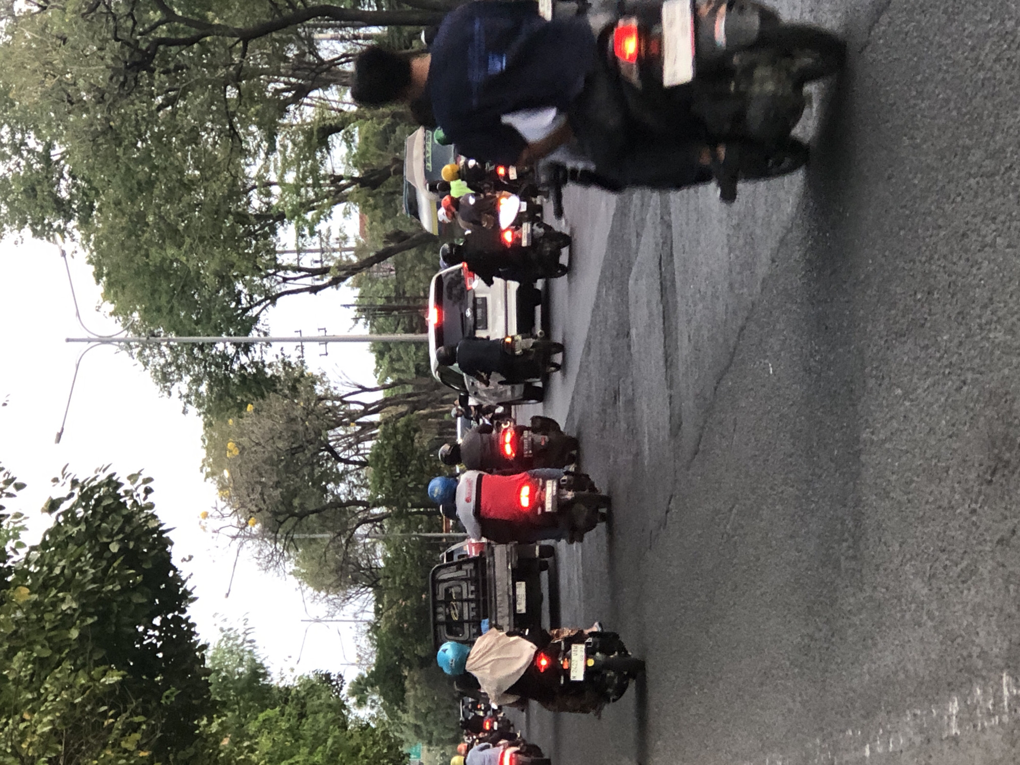The image depicts a traffic scene on a paved road in what appears to be a city in Indonesia, possibly Ngampelsari. The road is lined with trees on both sides, and there are streetlights visible. The sky is overcast.
The foreground is dominated by the road and several motorcycles with riders. A person in a dark blue shirt and black pants is riding a motorcycle in the right foreground, facing away from the camera. In the middle ground, several motorcycles are moving in the same direction as the foreground motorcycle. One motorcycle has a rider wearing a blue helmet and a beige scarf, and another has a rider in a red jacket with a blue helmet. Behind these are more motorcycles, a white SUV, and a gray pickup truck. Further in the background, there are more vehicles and trees. The general atmosphere suggests moderate traffic.