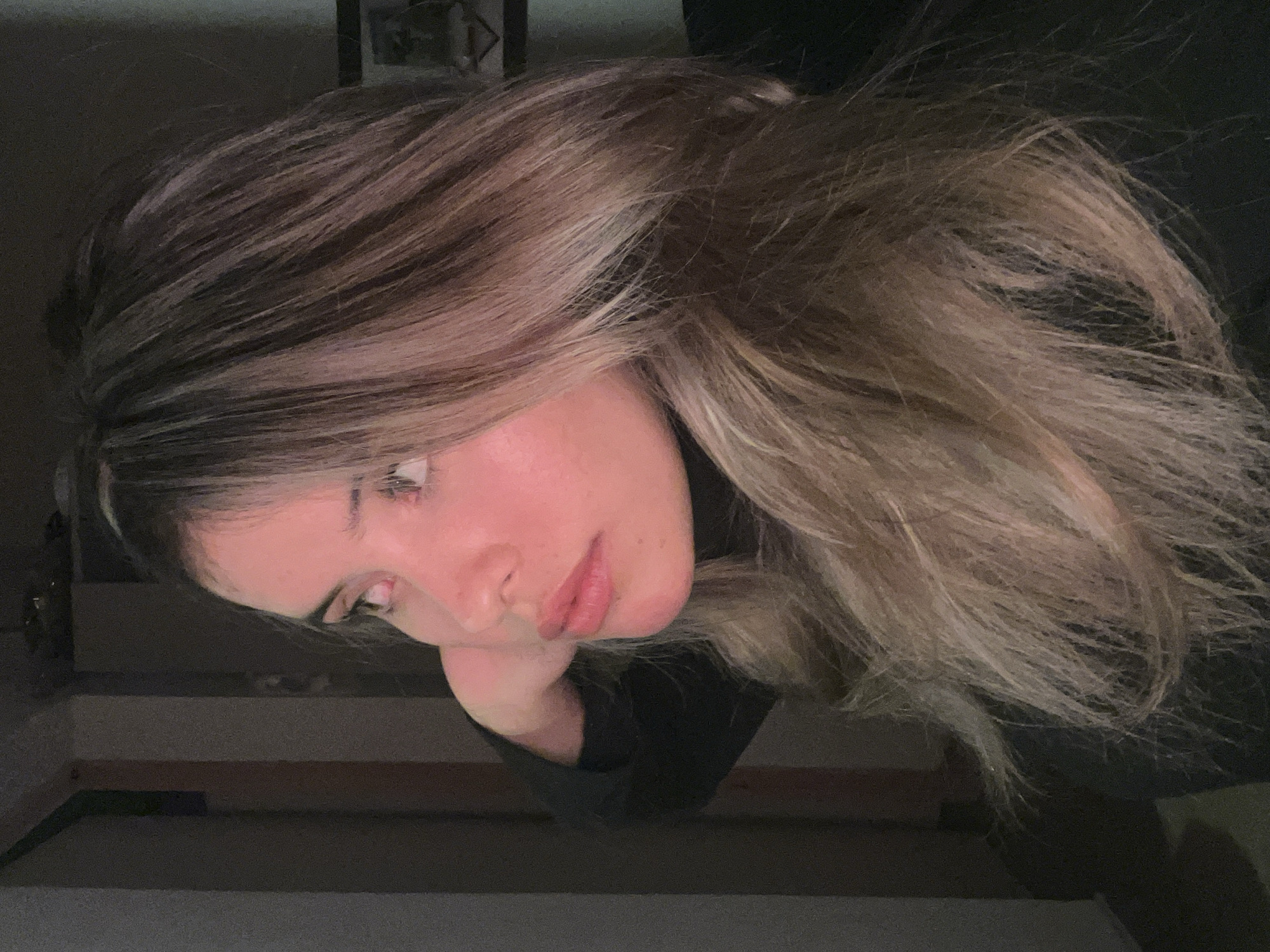 The image shows a close-up, profile view of a young woman with long, layered, blonde and brown hair. She is wearing a black turtleneck. Her right hand is resting behind her head. The lighting suggests an indoor setting, with a reddish hue cast over her face. The background is dark and indistinct, with some vertical elements that could be doorways or structural supports. There is a framed object visible in the upper right background. The scene does not depict any elements that can be used to identify a specific location in Sumalao, Argentina, or any other city.