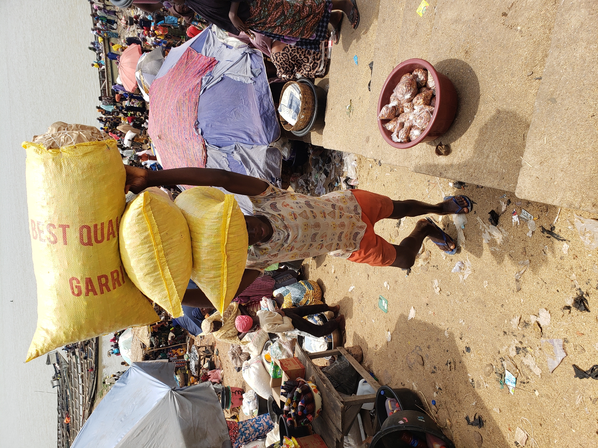 A sunlit outdoor market scene in Lokoja, Nigeria. A man in orange shorts and a patterned shirt carries two yellow sacks on his head, and a larger sack labeled "BEST QUALITY GARRI". Surrounding him are other vendors and customers amidst various goods and wares, including buckets of packaged goods, textiles, and baskets. The ground is littered with debris. In the background, a body of water is visible, with boats docked nearby. The scene suggests a bustling, informal trading environment.