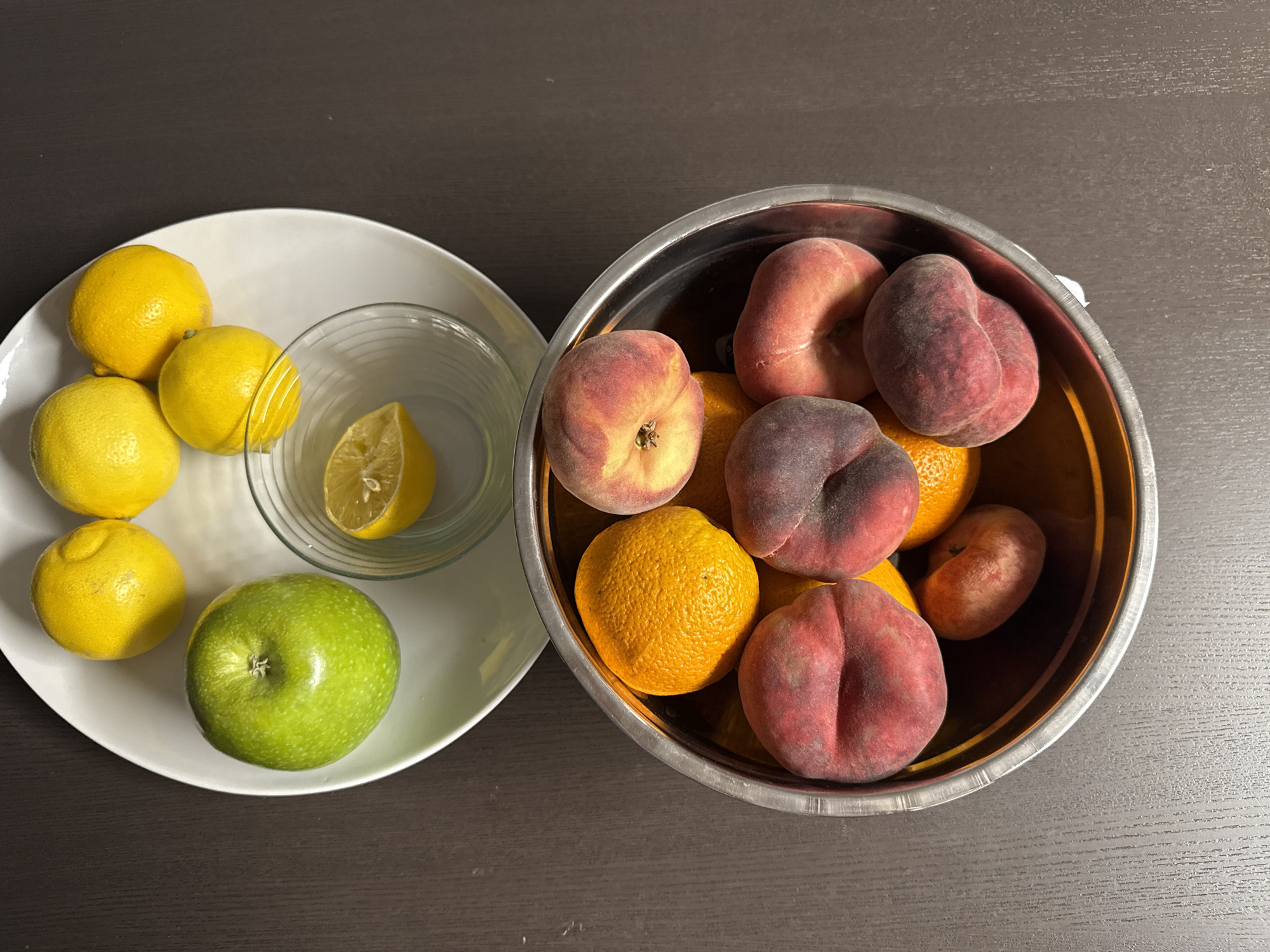 An overhead shot shows a collection of fruit arranged on a dark, wooden surface. In the upper portion of the image, a white circular platter holds several yellow lemons and one bright green apple. A small glass bowl containing a lemon half sits on the platter as well. Below the platter, a stainless steel bowl is filled with a mix of peaches and oranges. The peaches are predominantly red and yellow with some darker purple tones, and appear to be a variety with flattened sides. The oranges are a vibrant, deep orange. The lighting casts subtle shadows, highlighting the textures of the fruit and the wood grain of the surface. The scene is static and appears to be an indoor still life. This image does not contain any elements that would allow for a specific location in Puig-reig, Spain to be identified.