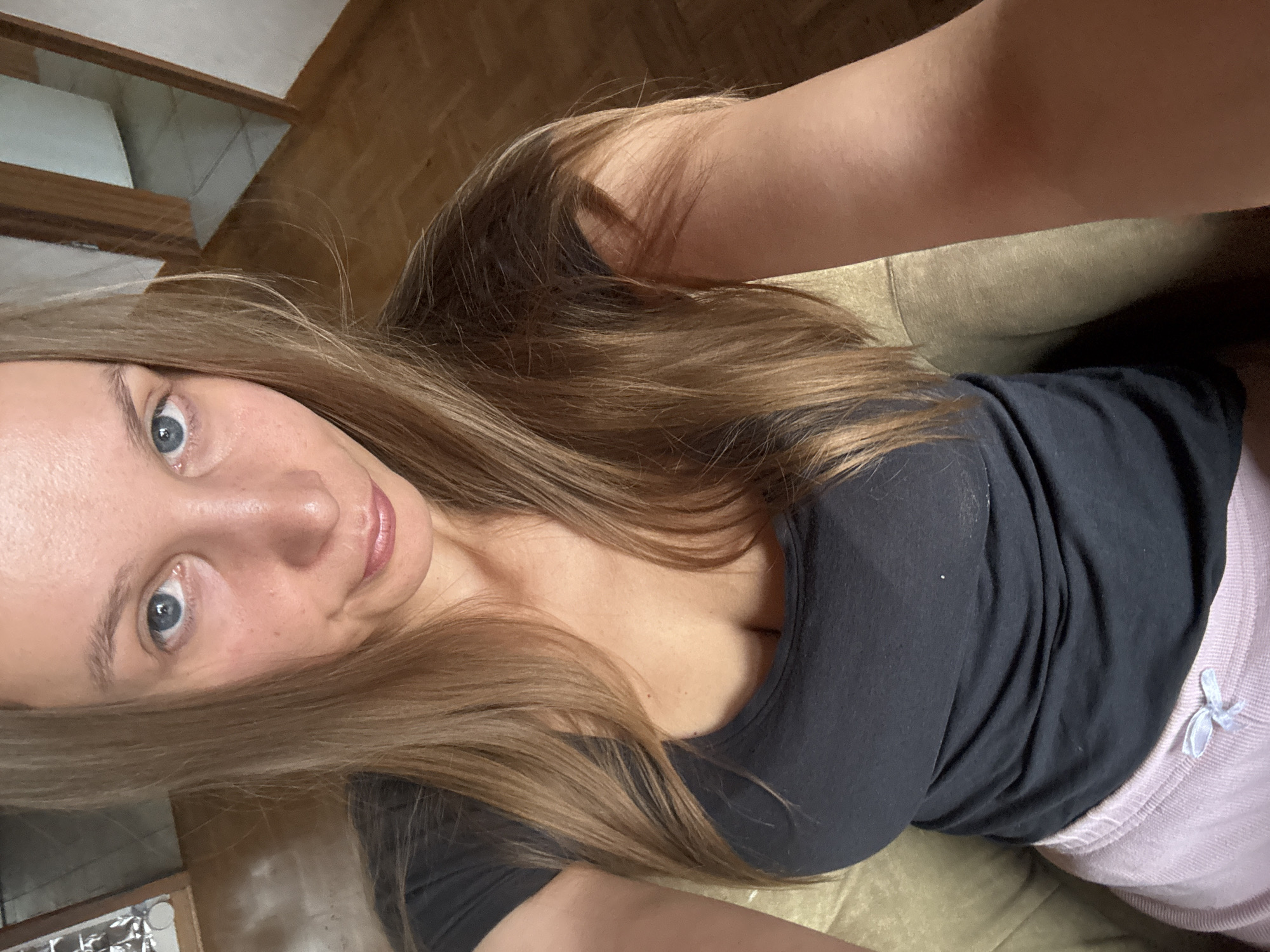 A woman is taking a selfie in a room that appears to be a living room. She is sitting on a sofa or chair, wearing a black t-shirt and pink pajama bottoms. Her hair is long and blonde, and she has blue eyes. The room has wooden floors and a doorway leading to another room. There is furniture visible in the background. The location is identified as Überherrn, Germany.