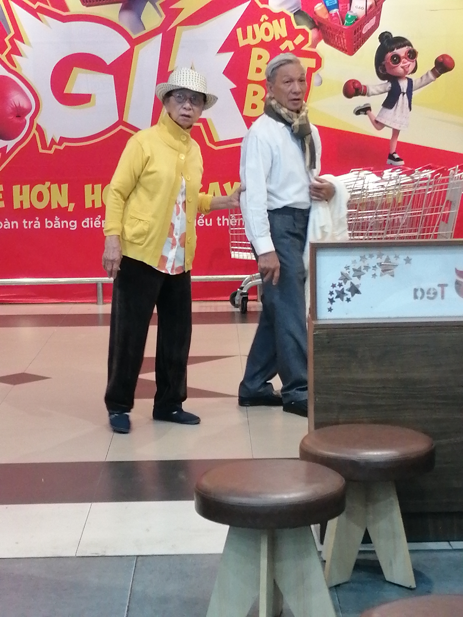 Two elderly individuals, a woman and a man, are standing in what appears to be an indoor market or commercial space in Da Lat, Vietnam. The woman, on the left, is wearing a yellow cardigan over a patterned shirt and dark pants. She has on a straw hat and glasses. The man, on the right, is wearing a white shirt, a scarf, and dark pants. He is holding a white cloth. They are both looking in different directions. Behind them, a large red banner with text and images is displayed, including cartoon characters and boxing gloves. A shopping cart is also visible. The flooring appears to be tiled with a geometric pattern. In the foreground, two small wooden stools with brown seats are present.