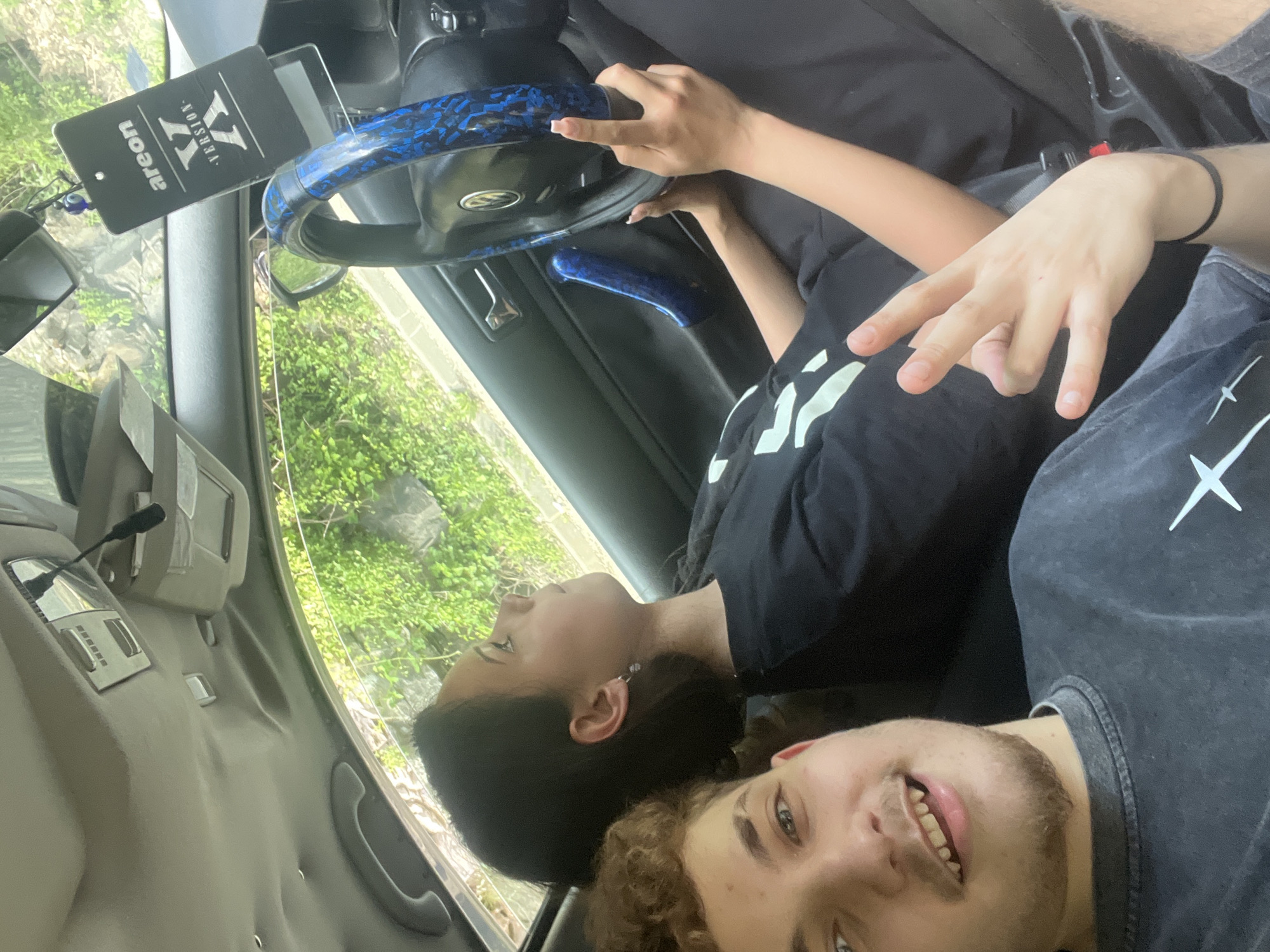 Two young individuals, a male and a female, are positioned inside a car. The male is in the foreground, looking towards the camera with a smile, his face partially visible. He is wearing a dark-colored t-shirt with a white graphic. The female is seated in the driver's seat, looking away from the camera and towards the right. She has dark hair and is wearing a black t-shirt. Her hands are on the steering wheel, which is covered with a blue patterned accessory. A black air freshener is hanging from the rearview mirror. The interior of the car, including the dashboard and door panel, is visible. Outside the car, lush green foliage suggests an outdoor environment, possibly a roadside or park. The image appears to be taken in Vlahovo, Bulgaria, based on the contextual information provided.